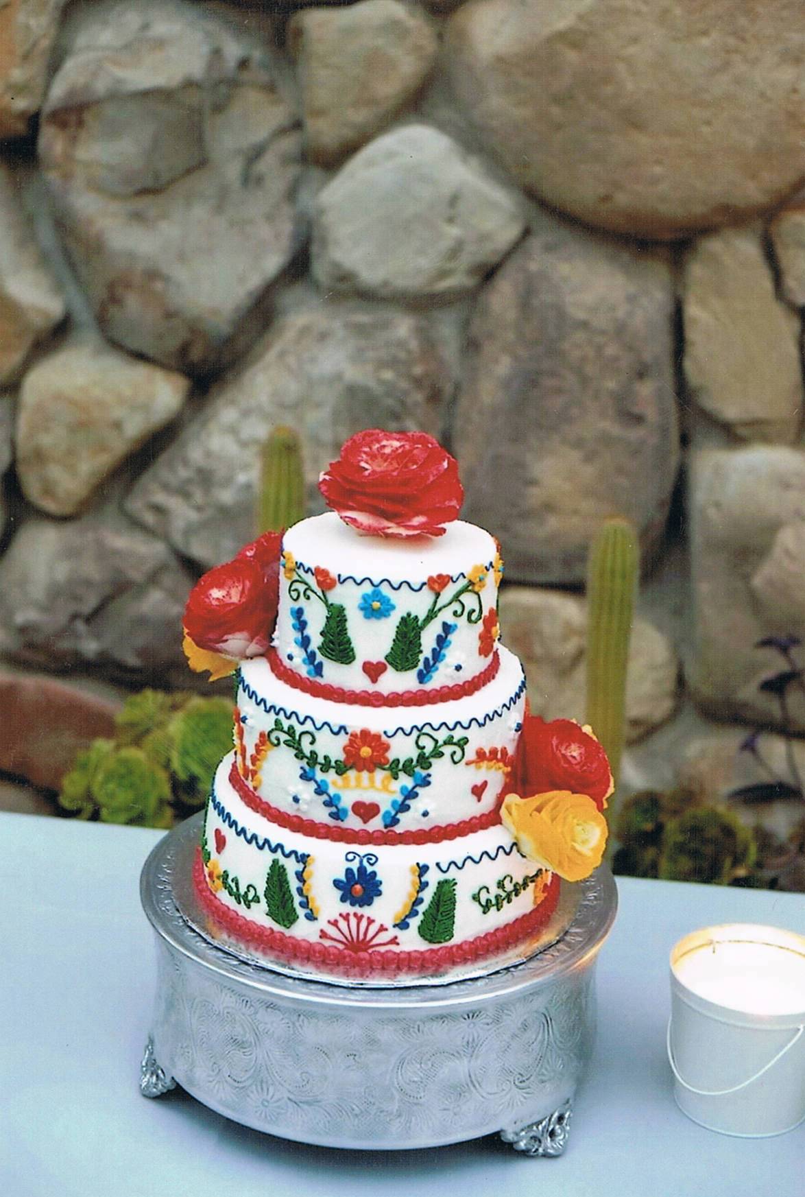 Three-tiered cake decorated with colorful floral and cactus patterns, topped with a red flower, against a stone wall background.