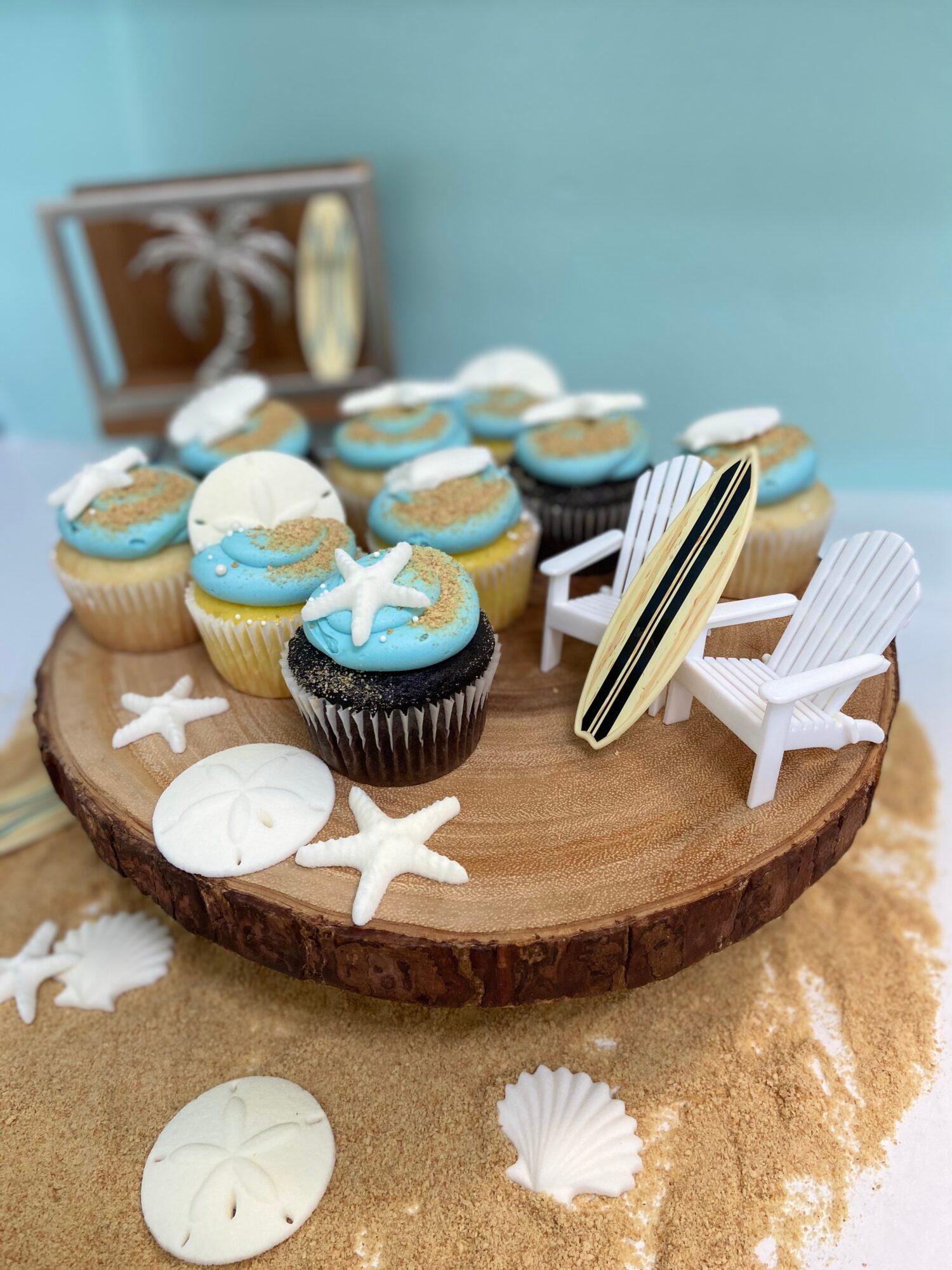 Cupcakes with blue and white frosting, starfish decorations, miniature beach chairs, and surfboard on a wooden cake stand, beach-themed setup.