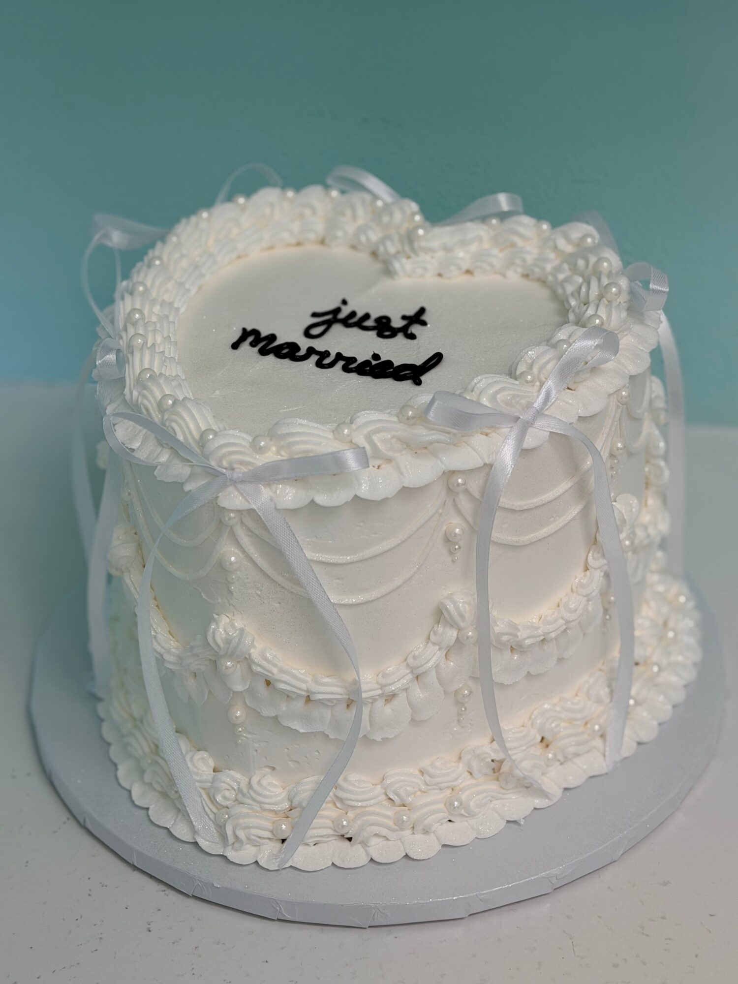 Round wedding cake with white icing, decorative ribbons, and a message on top, on a white base.