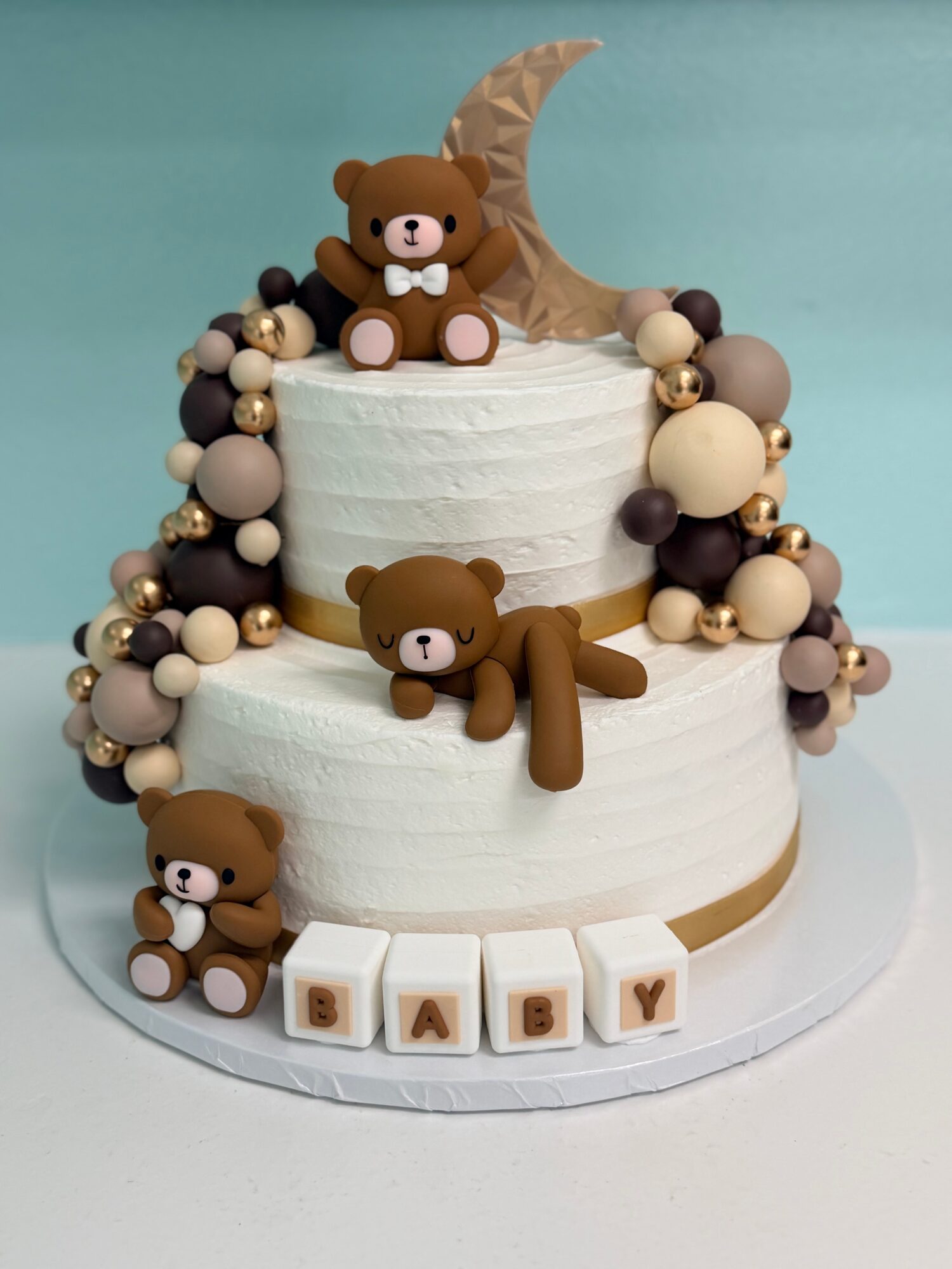 Two-tiered white cake decorated with teddy bear figures, beads, and blocks spelling 'BABY'.