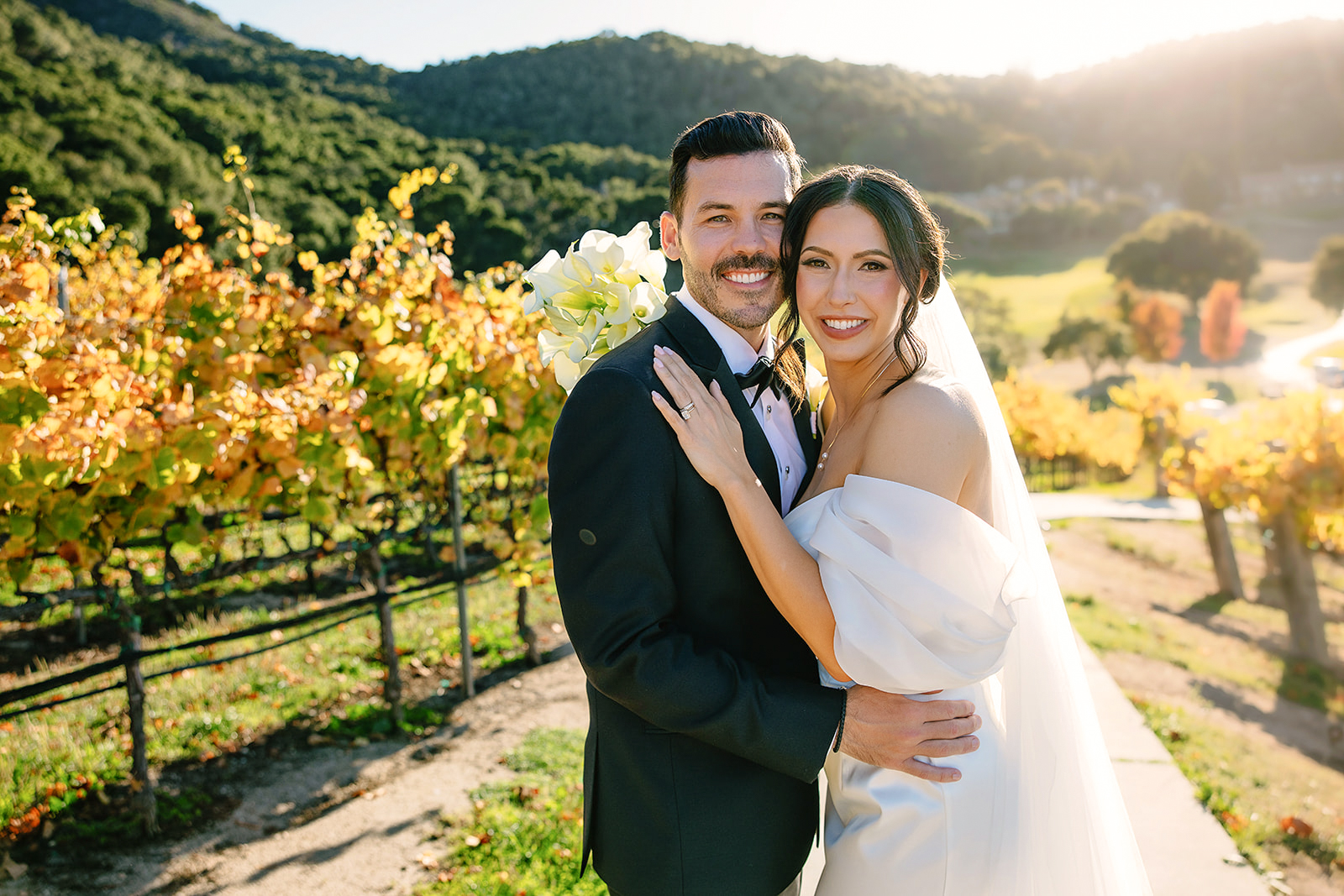A smiling couple in wedding attire stands outdoors in a vineyard during sunset, with trees and hills in the background.