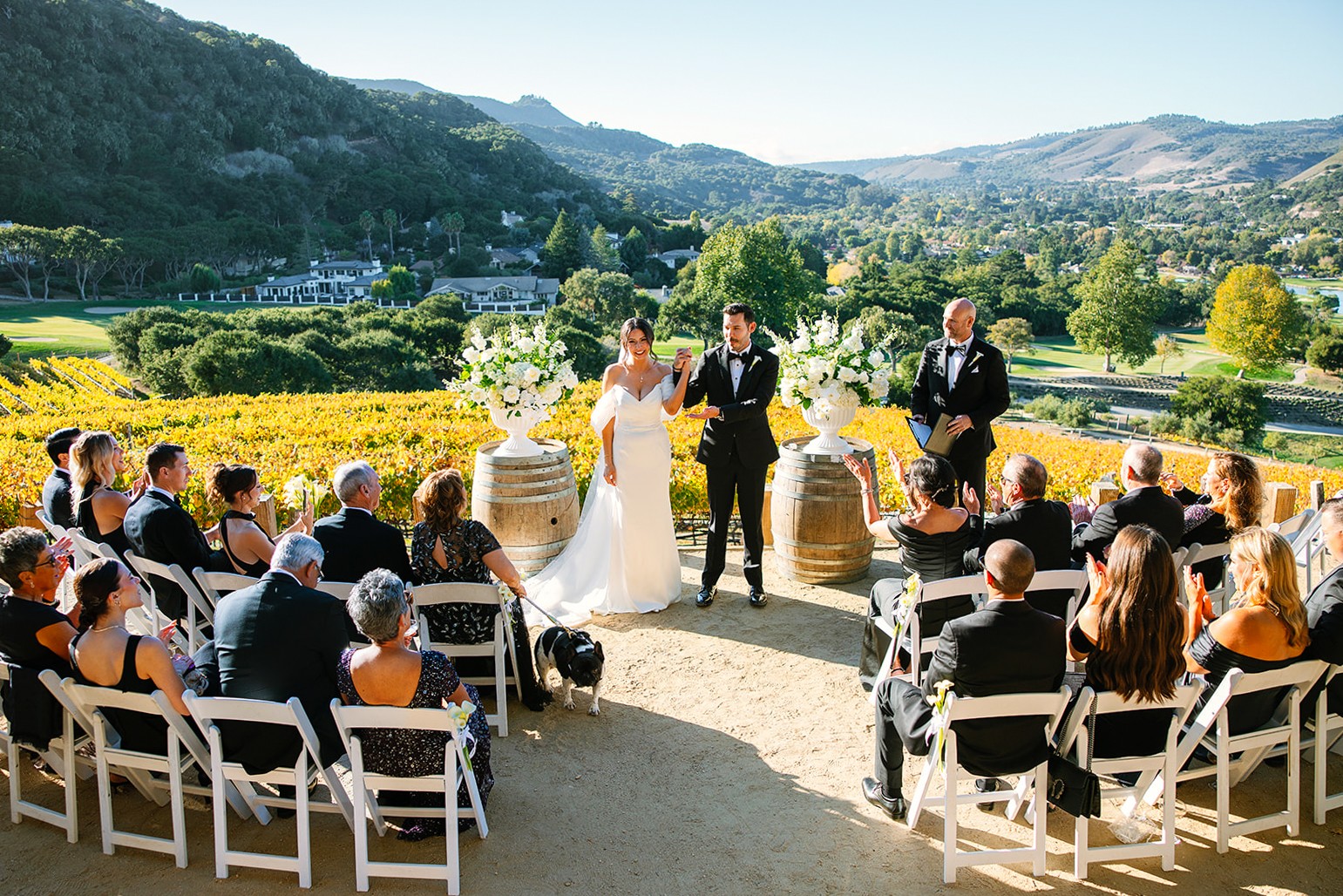 A wedding ceremony outdoors with a bride, groom, and officiant standing before seated guests, scenic landscape background.