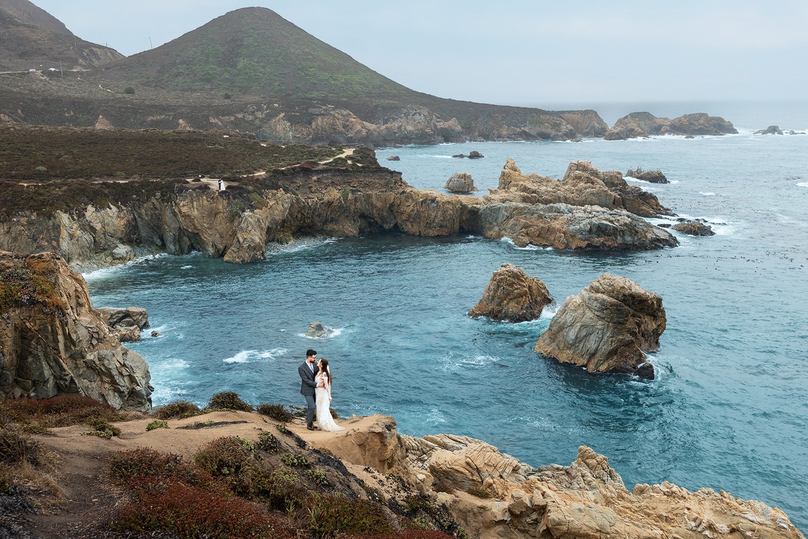 Couple standing on rocky coast near ocean with cliffs and hills in background.