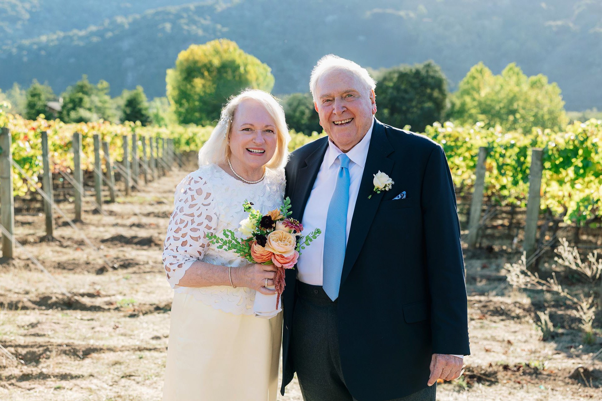 Two smiling elderly people, a woman holding a bouquet, standing in a vineyard with green trees and mountains in the background.