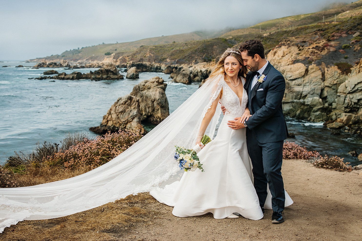 Bride and groom standing on a rocky coastline, embracing, with the bride holding a bouquet, ocean and cliffs in background.