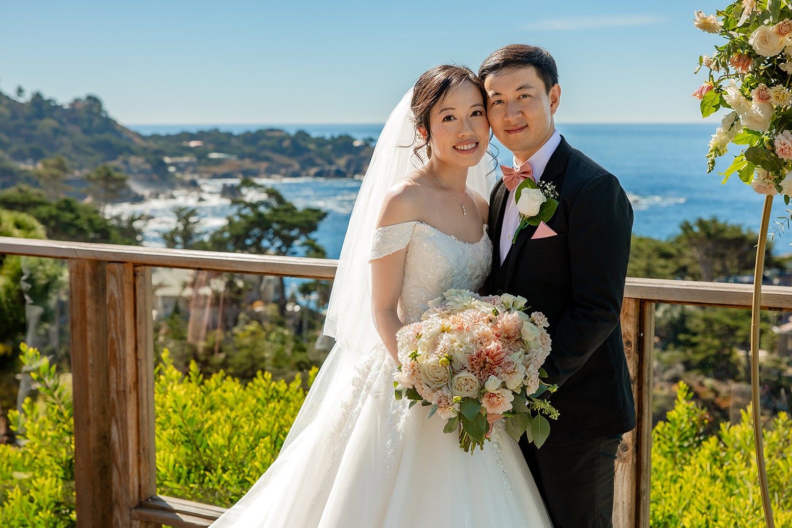 Bride and groom smiling outdoors with ocean and landscape in background, bride holding a bouquet, wedding attire, sunny day.