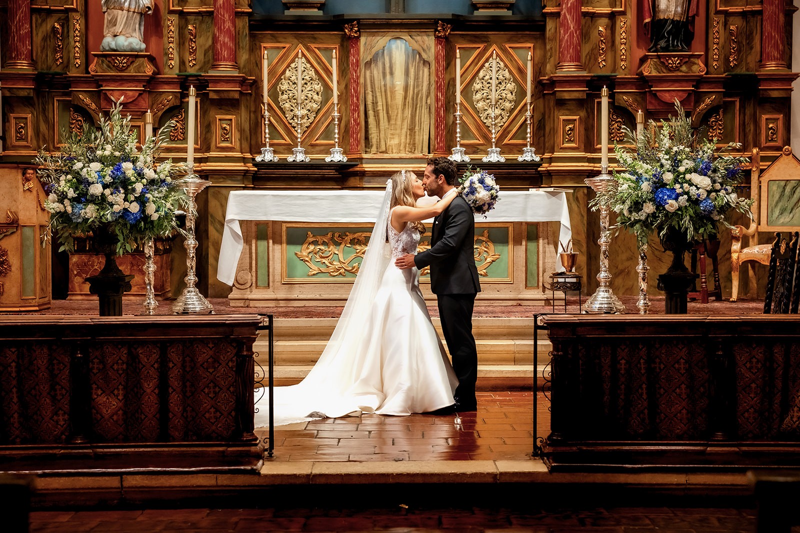Couple dancing at wedding altar with floral arrangements and ornate wooden backdrop, in a church setting.