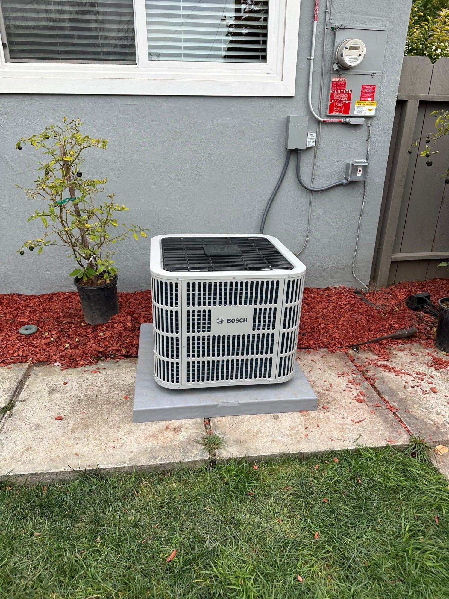 Outdoor air conditioning unit on a concrete pad next to a gray wall with a window and electrical connections, with a small plant nearby.