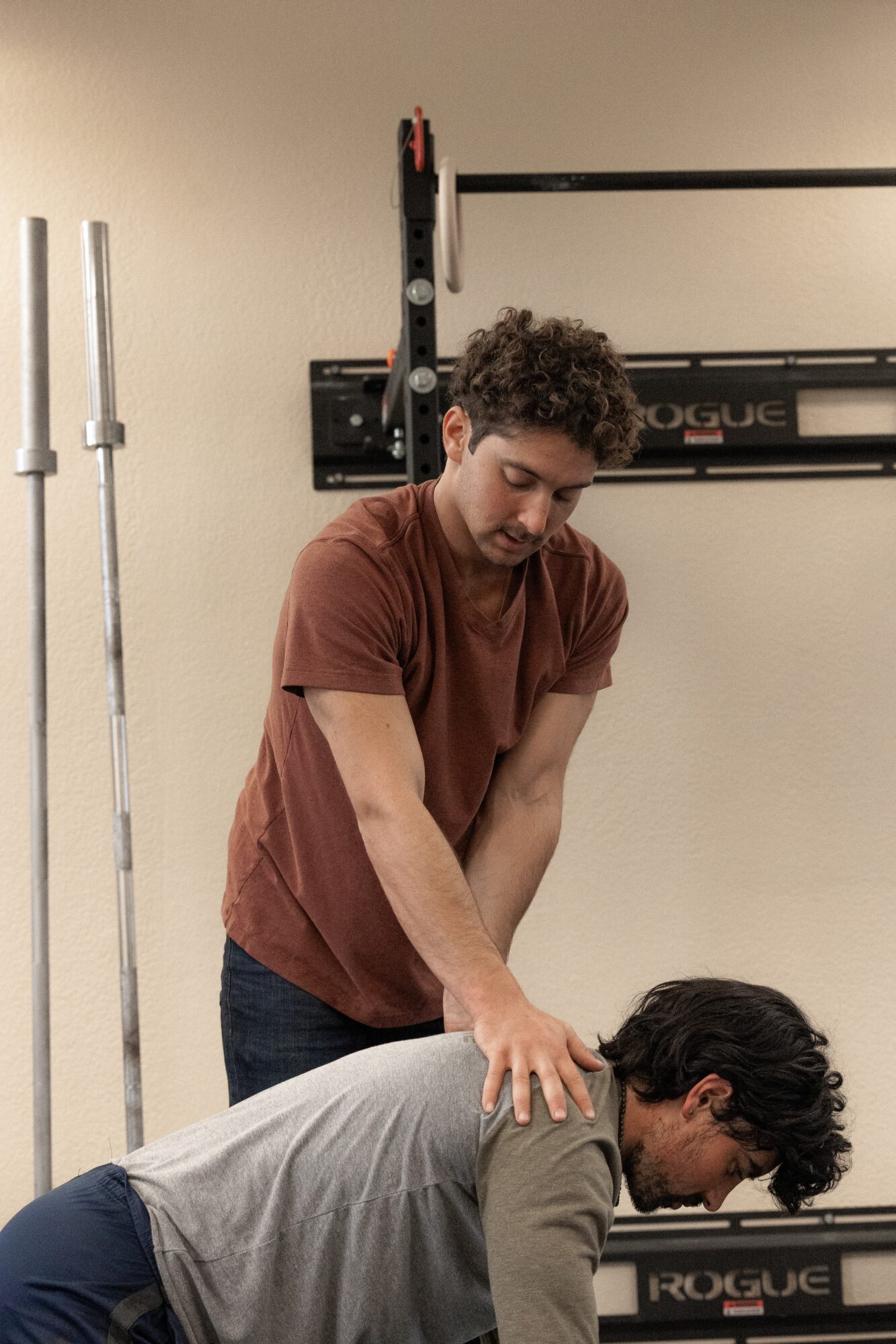 Person in gray shirt receiving shoulder massage from person in brown shirt in gym setting.