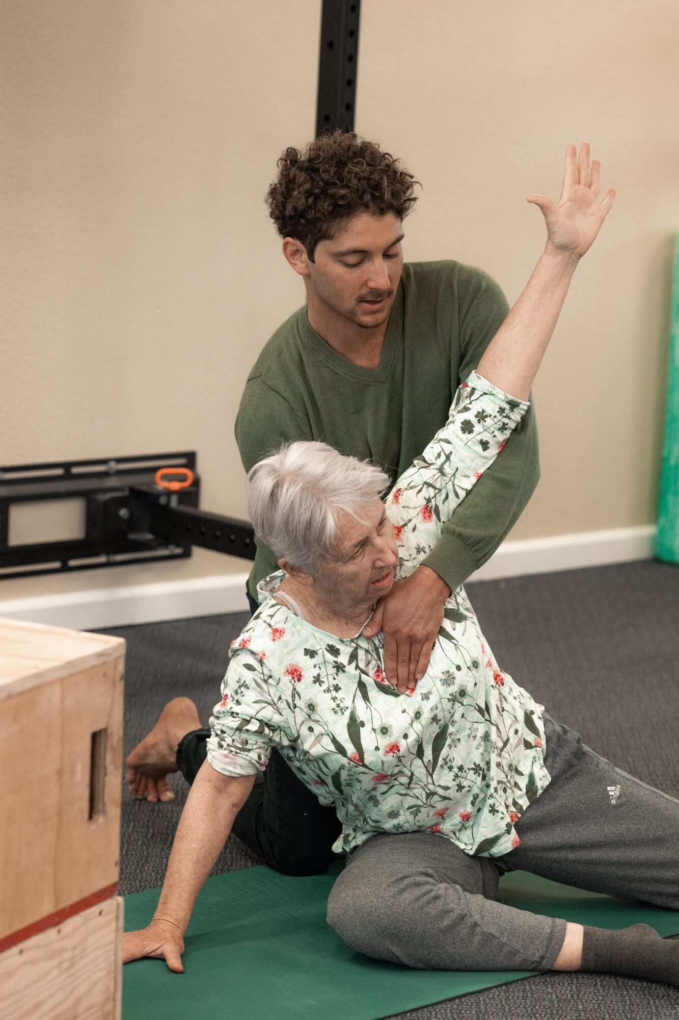 Young man assisting an elderly woman in a seated stretch, raising her arm, in a room with a green mat and beige wall.