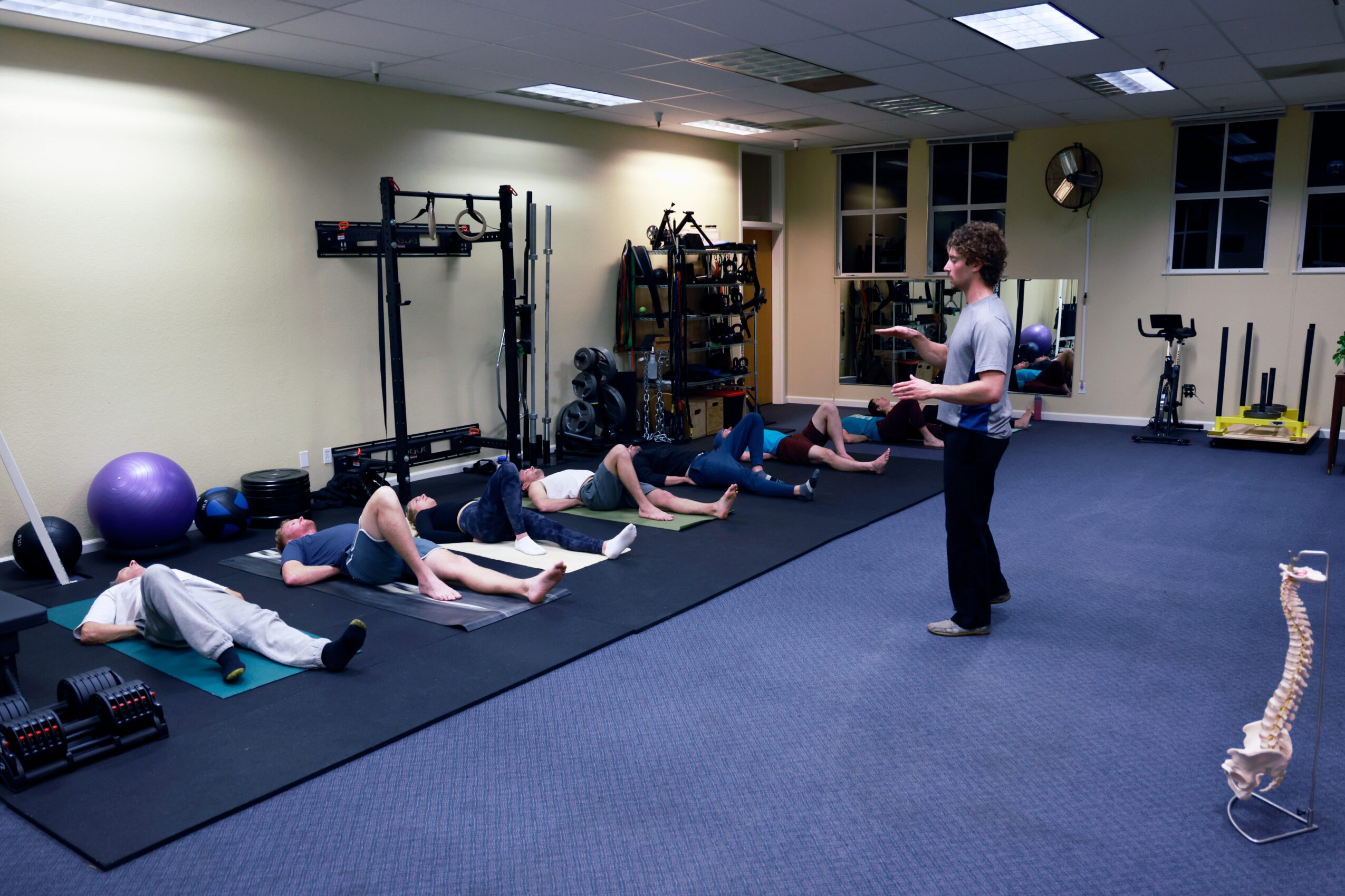 Instructor standing in fitness room with five people lying on mats, stretching legs, exercise equipment in background, skeleton model nearby.