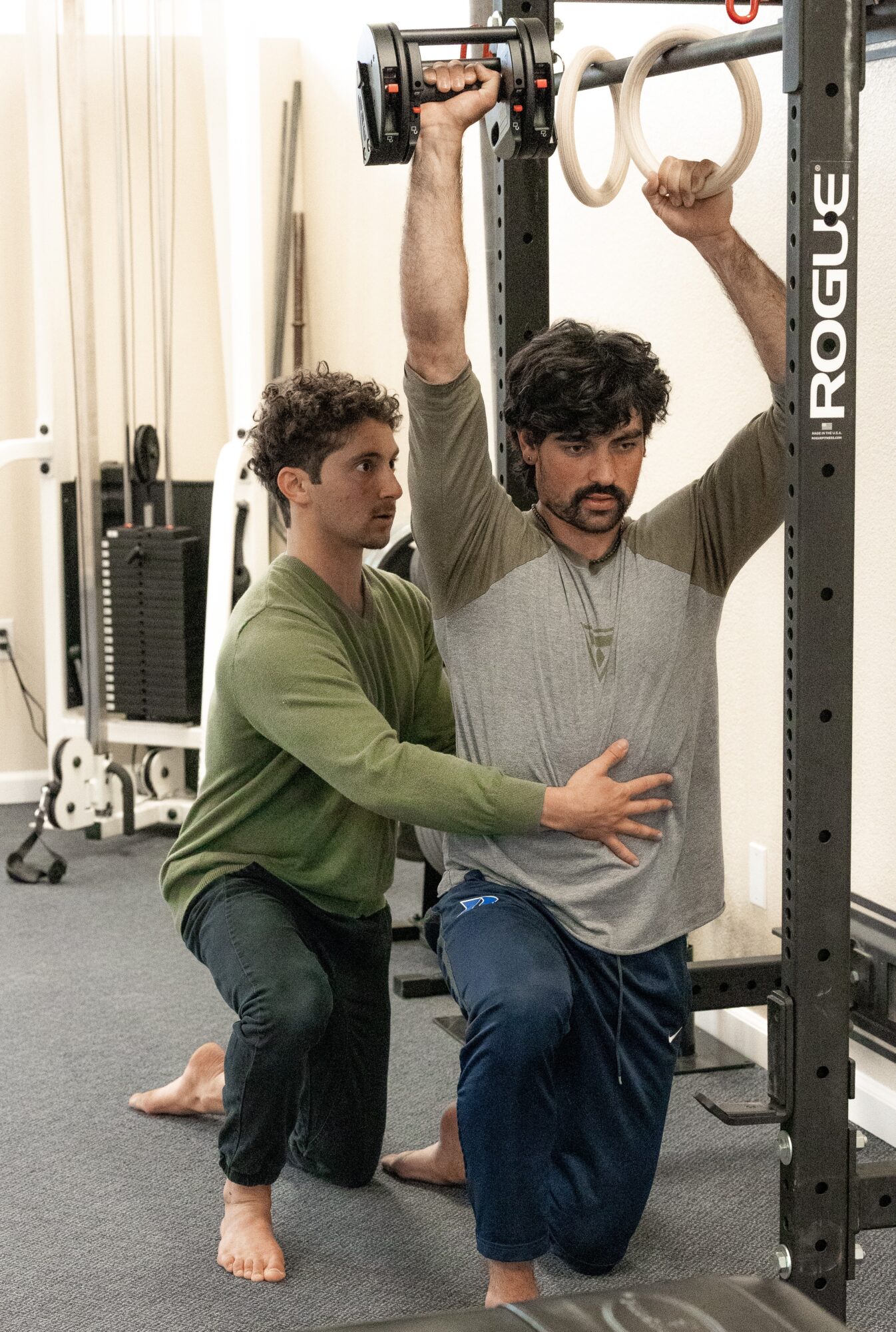 Two men in a gym, one assisting the other with gym rings, both kneeling on the floor, equipment in background.