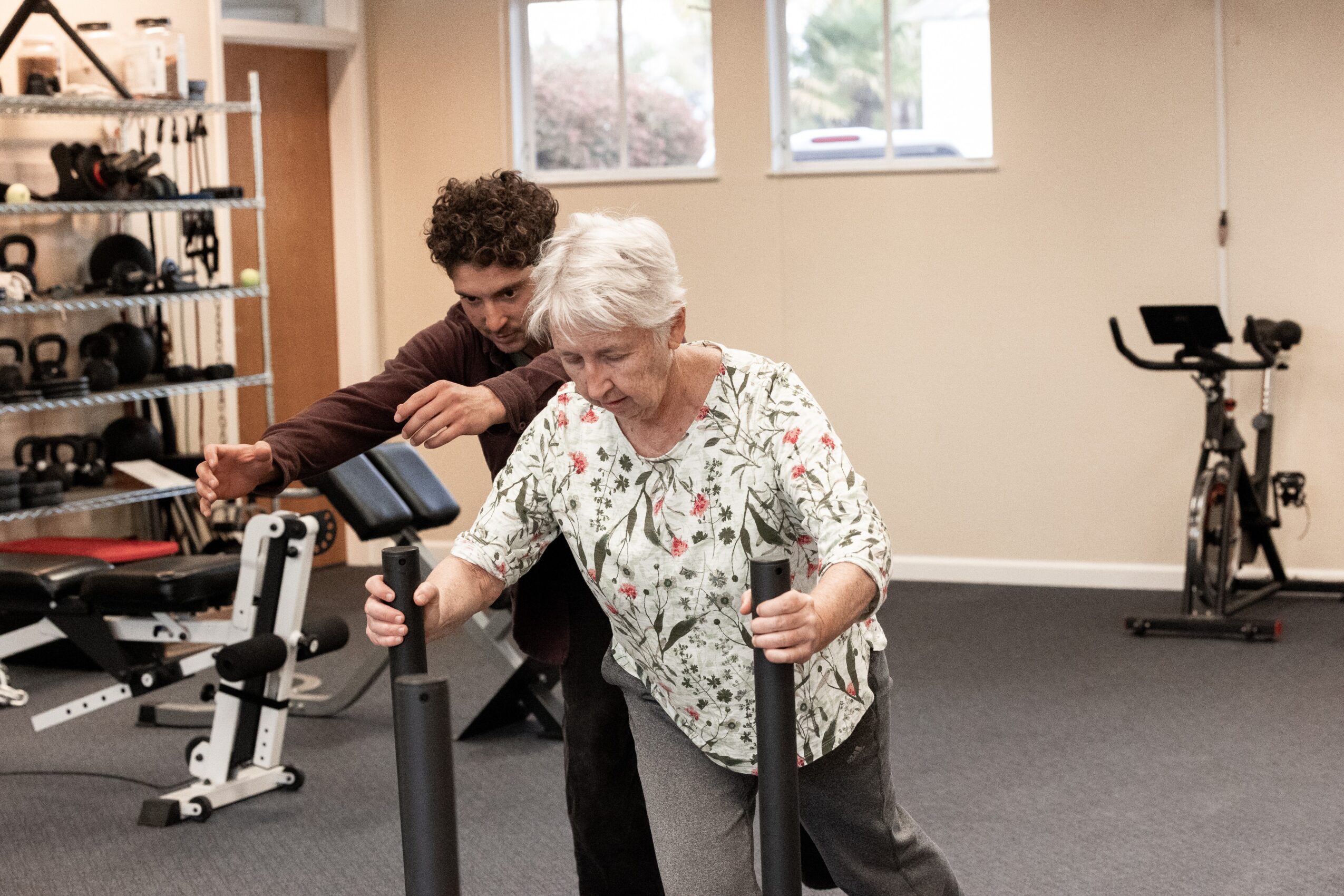 Older woman and young man exercising with stationary bikes in a gym, with fitness equipment in the background.