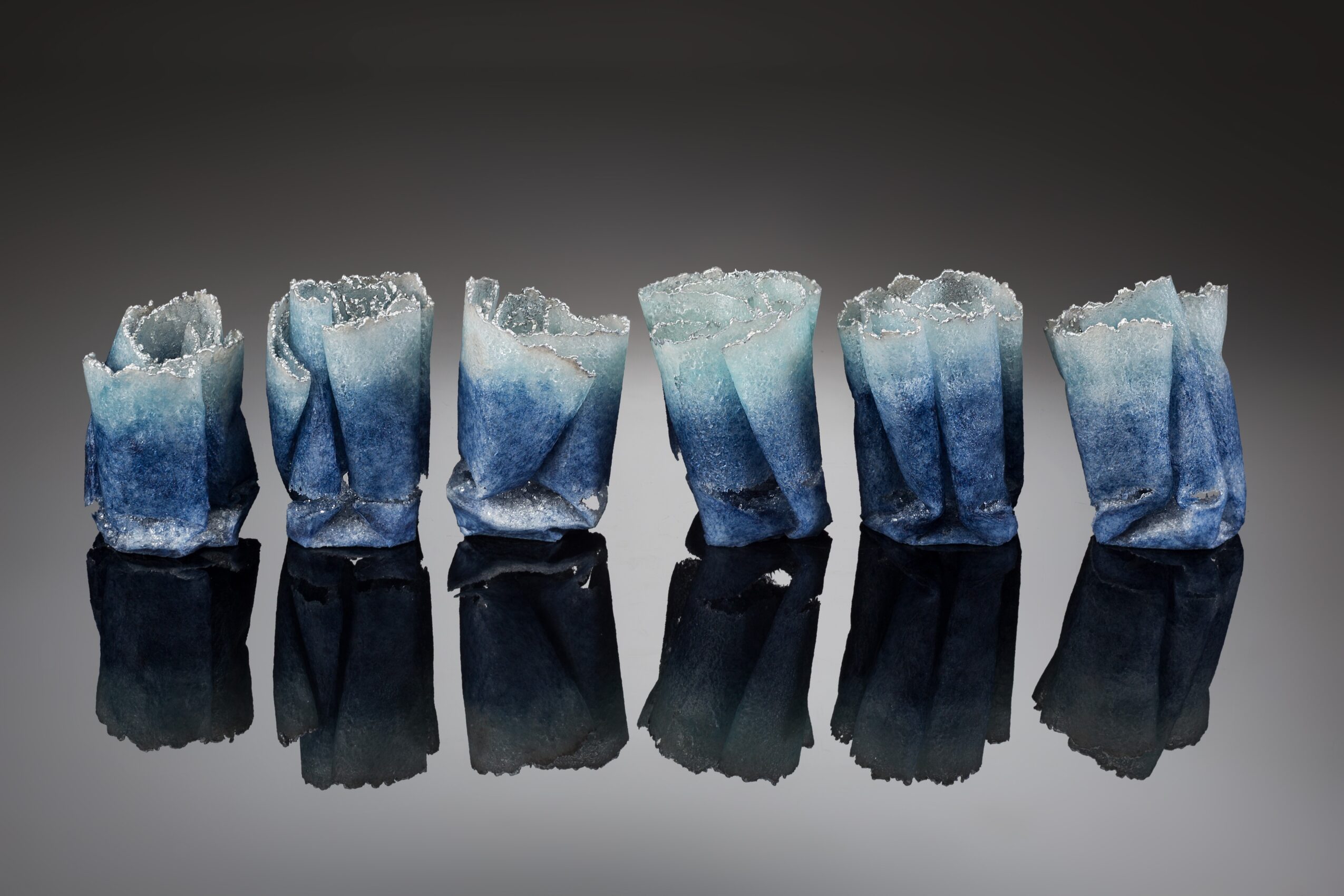 Six crumpled blue and white paper cups are arranged in a row on a reflective black surface, with a dark background.