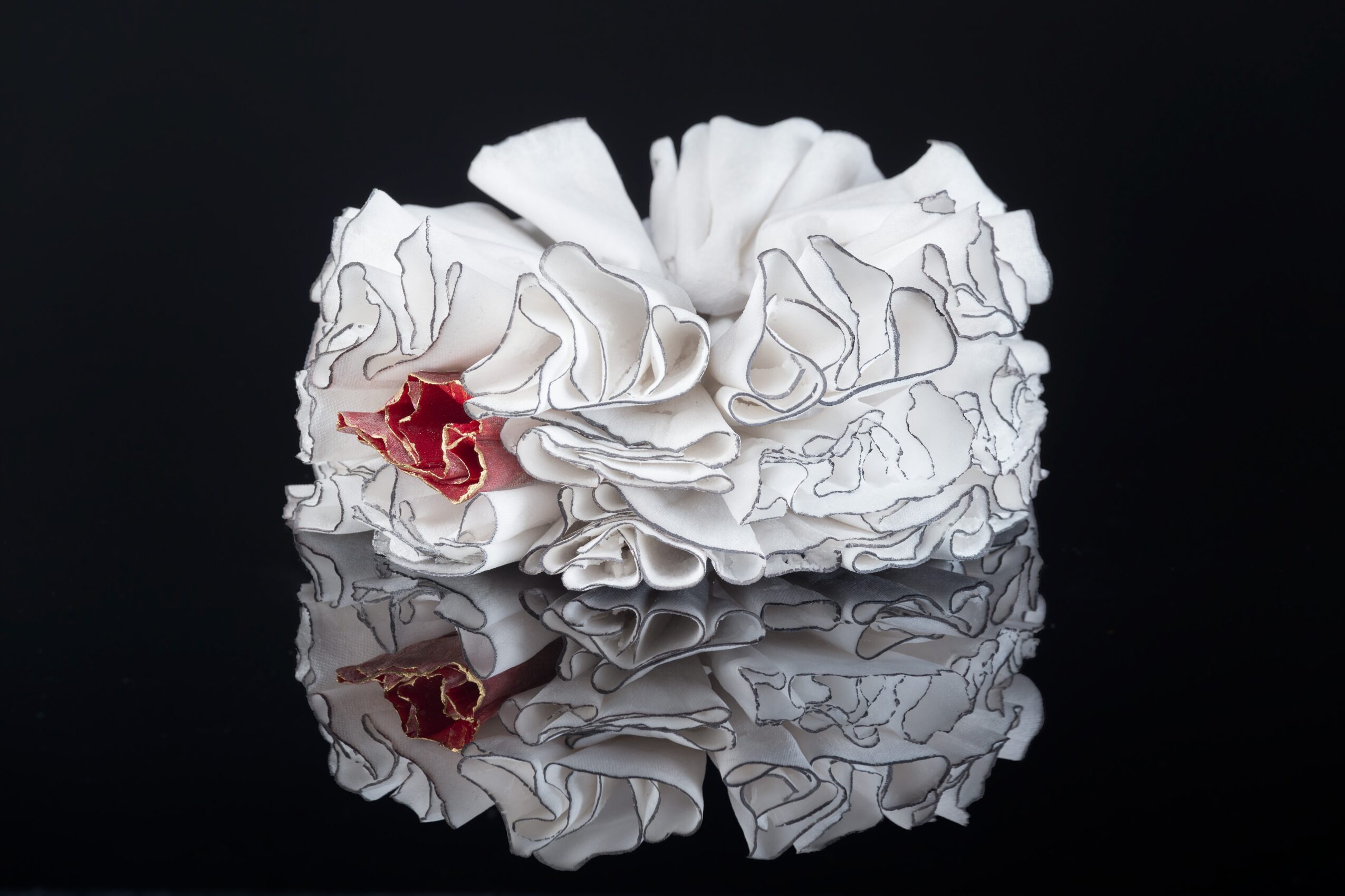 White ruffled fabric with a small red flower, reflected on a shiny surface, against a black background.