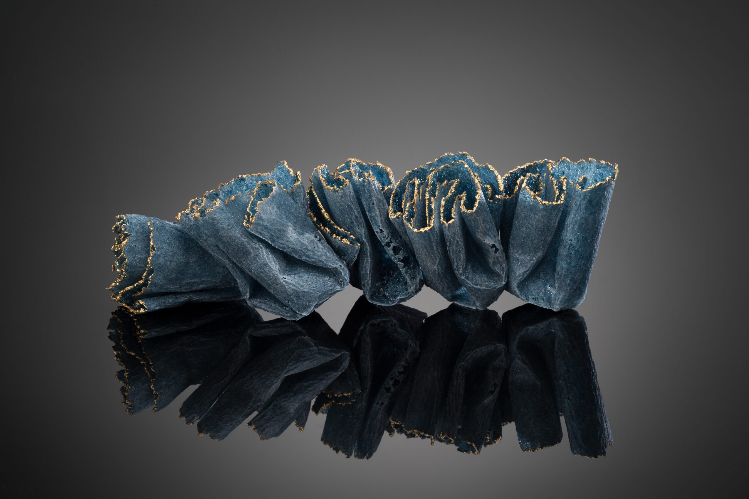 Blue fabric scrunchie with gold embroidery, reflected on a glossy surface, against a dark background.