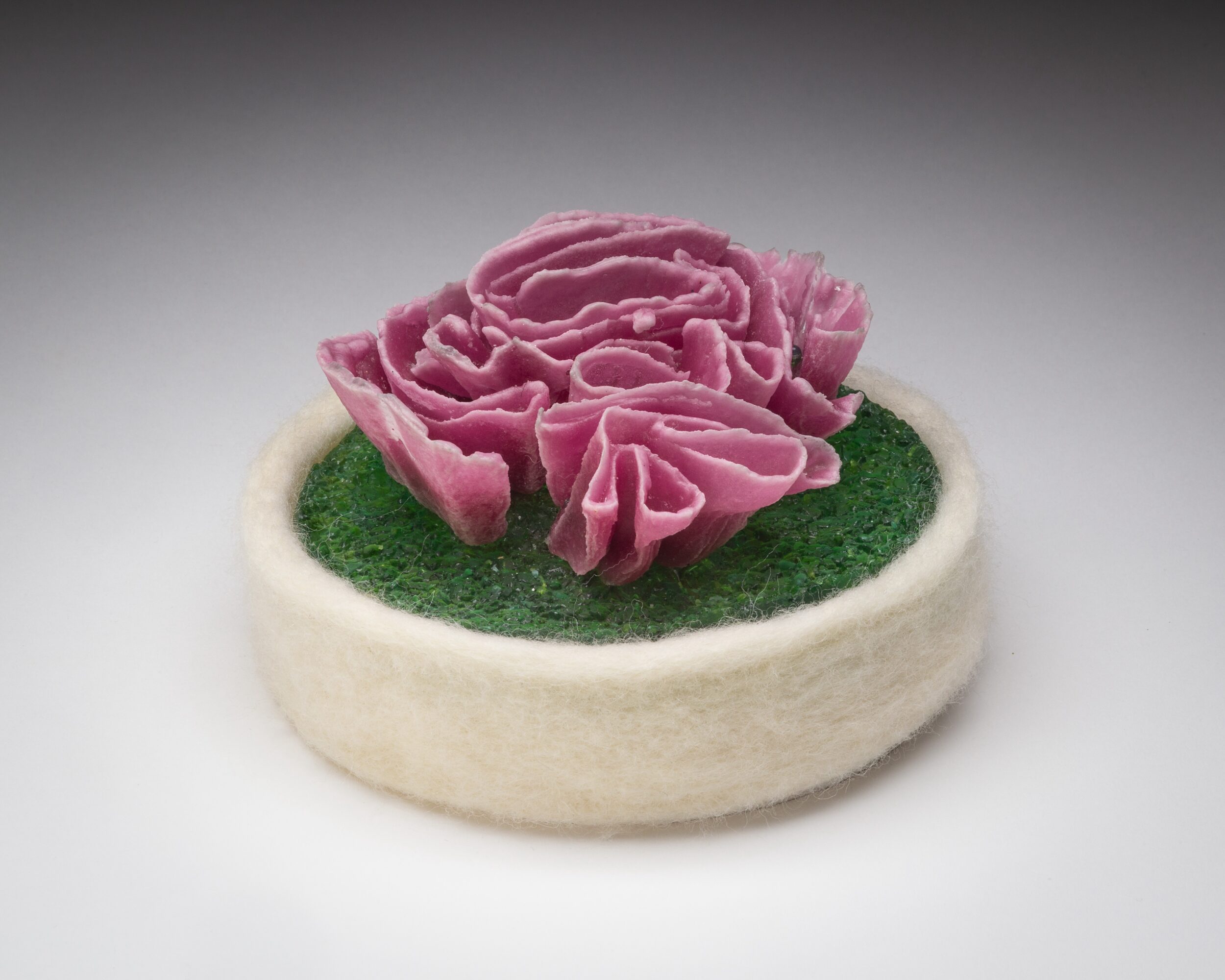 A small round object with a white base, green surface, and pink flower on top, against a plain background.