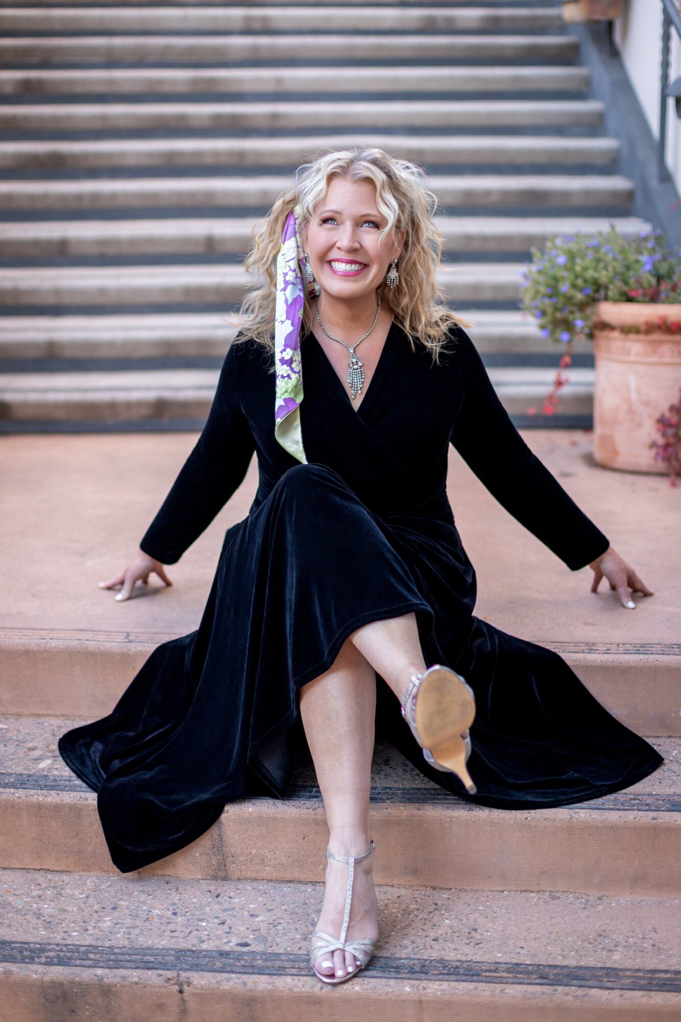Woman with curly blonde hair sitting on stairs, smiling, wearing a black dress and high heels, with potted plants nearby.