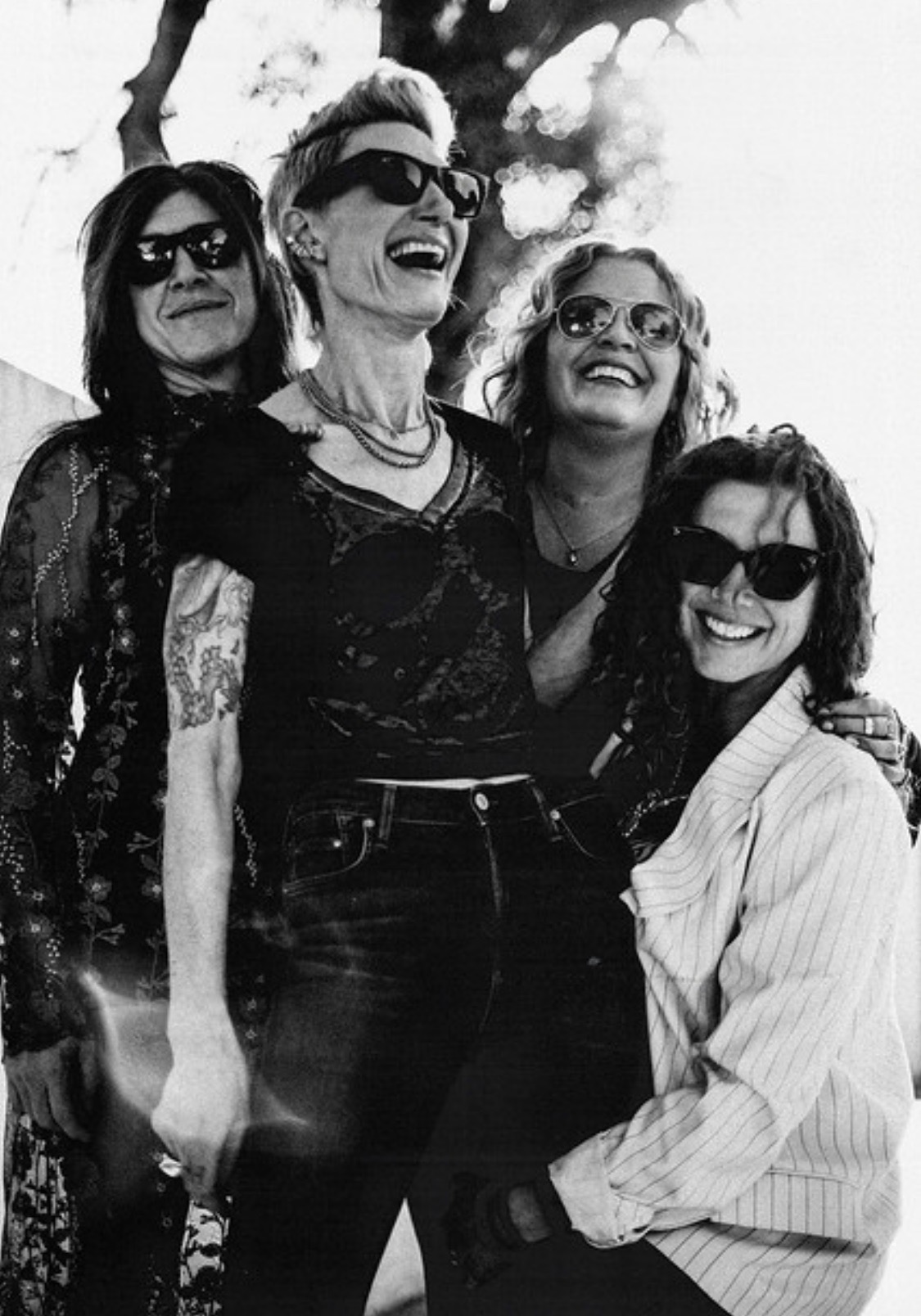 Four women smiling and wearing sunglasses outdoors, standing close together, with trees in the background.
