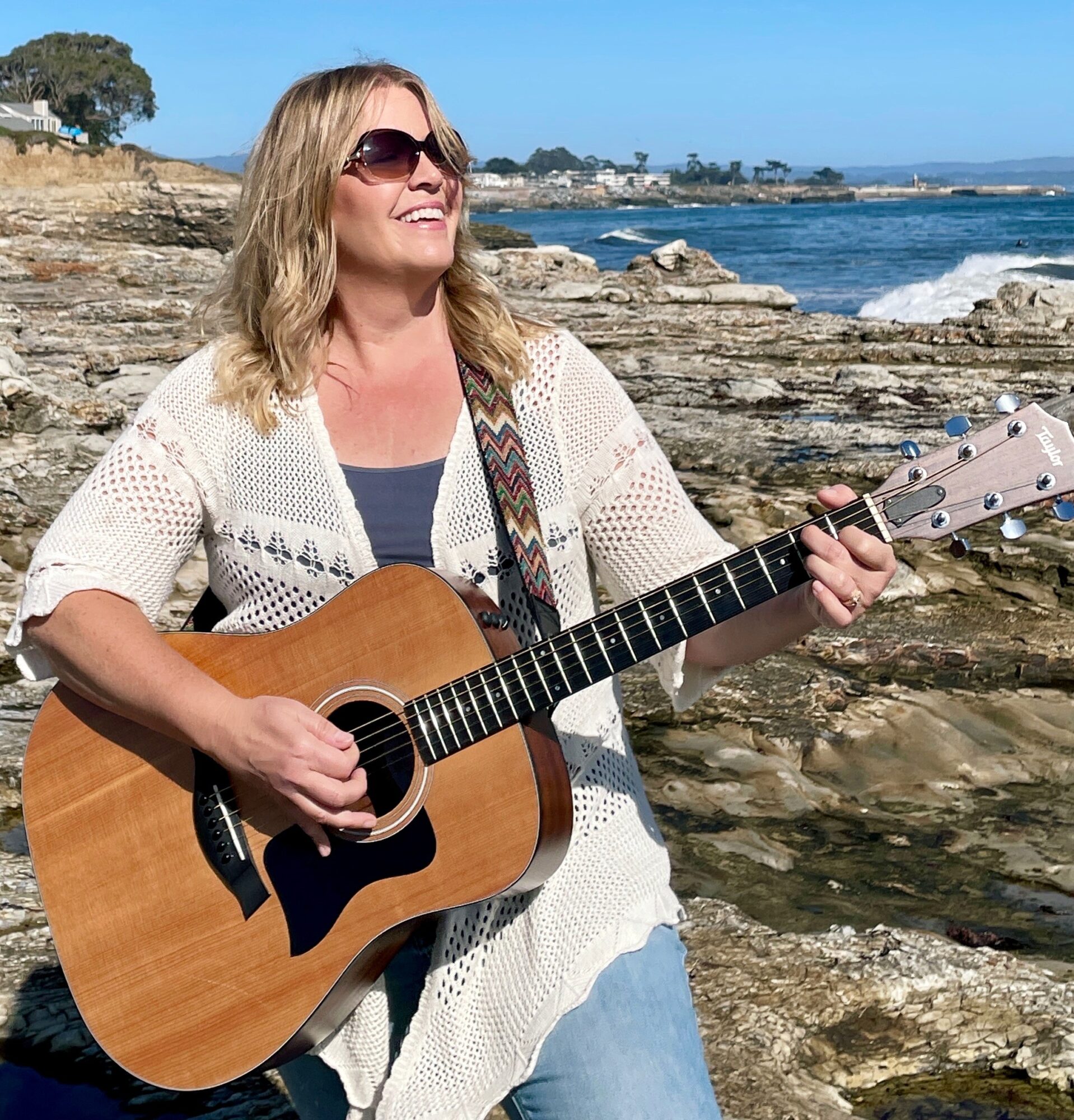 Woman playing guitar on rocky shoreline with ocean and blue sky in background.