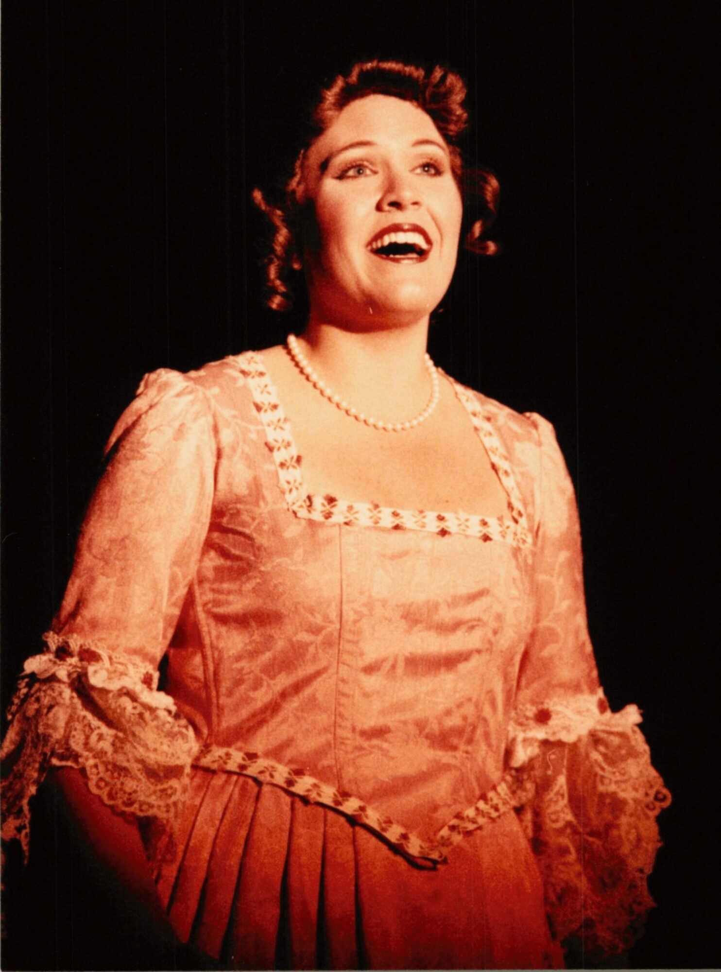 Woman singing or performing on stage, wearing a vintage-style dress with lace details and a pearl necklace.