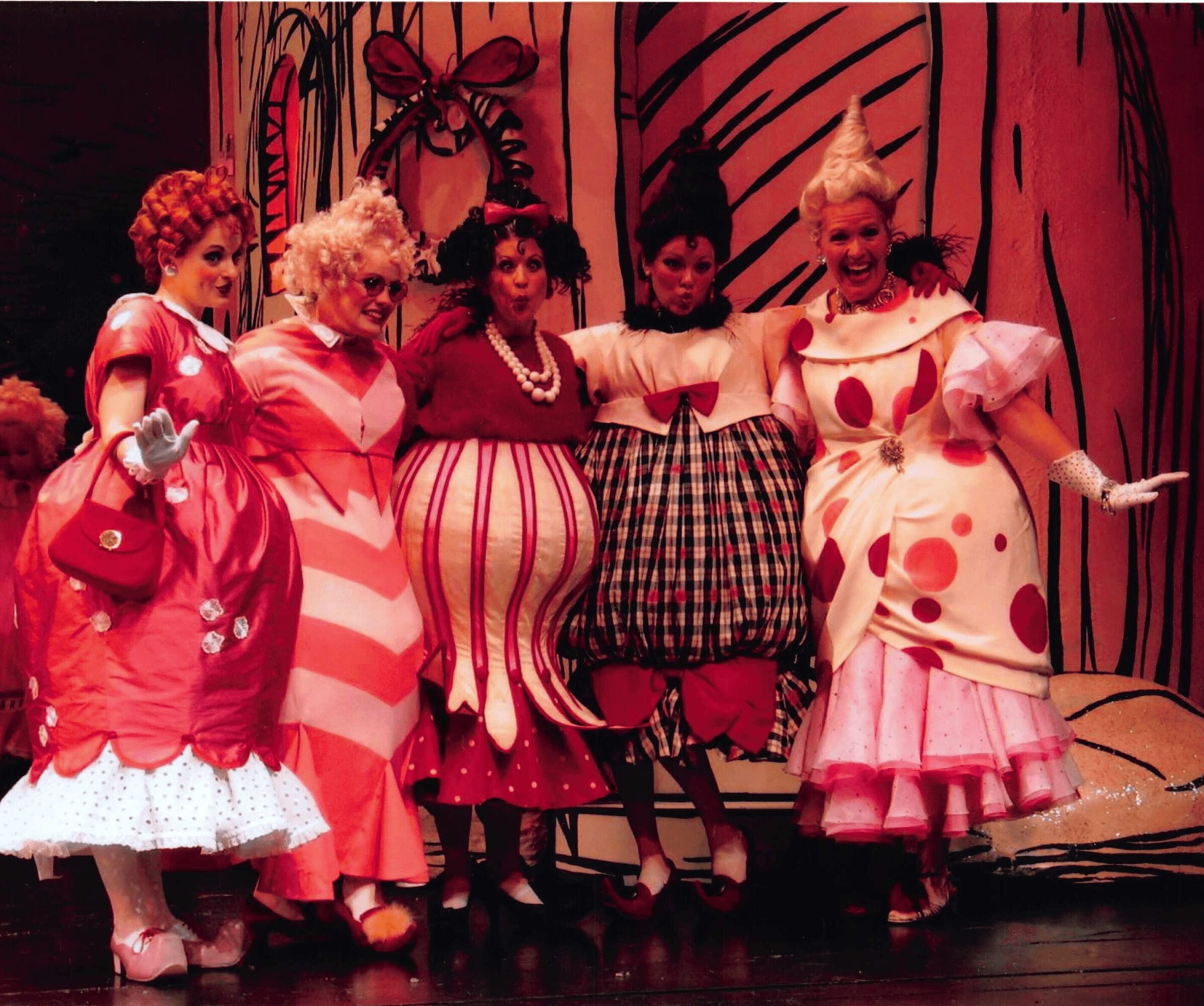 Group of women in colorful, whimsical costumes on stage with a decorated background, smiling and posing together.