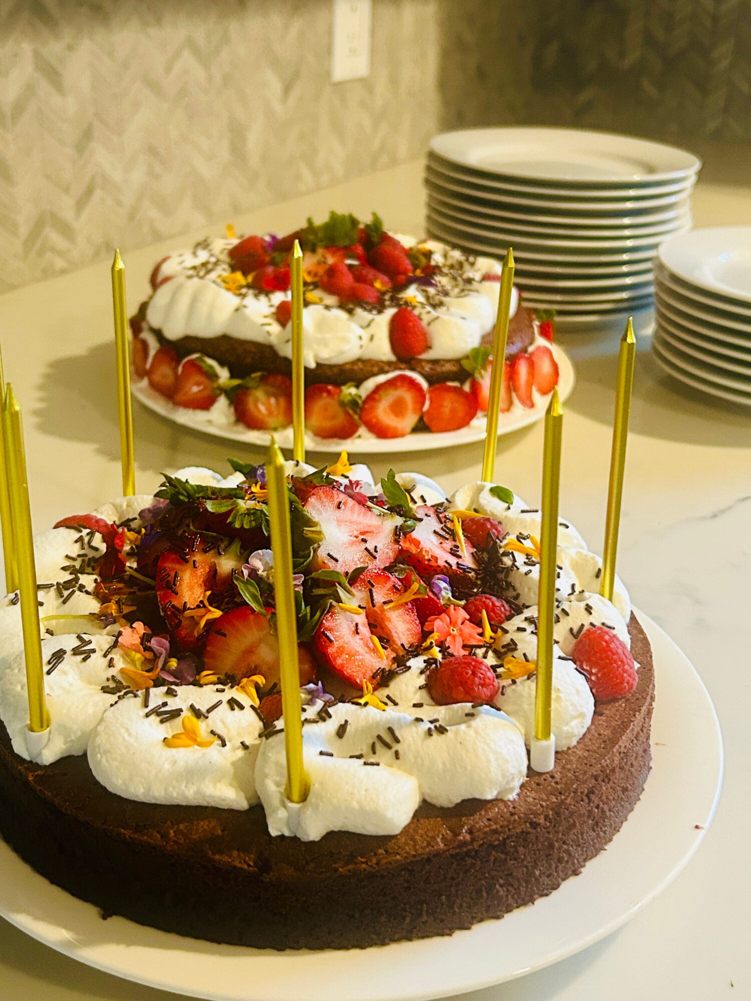 Two-layer cake with strawberries, whipped cream, and yellow candles, decorated with fruit and chocolate, on a white plate.