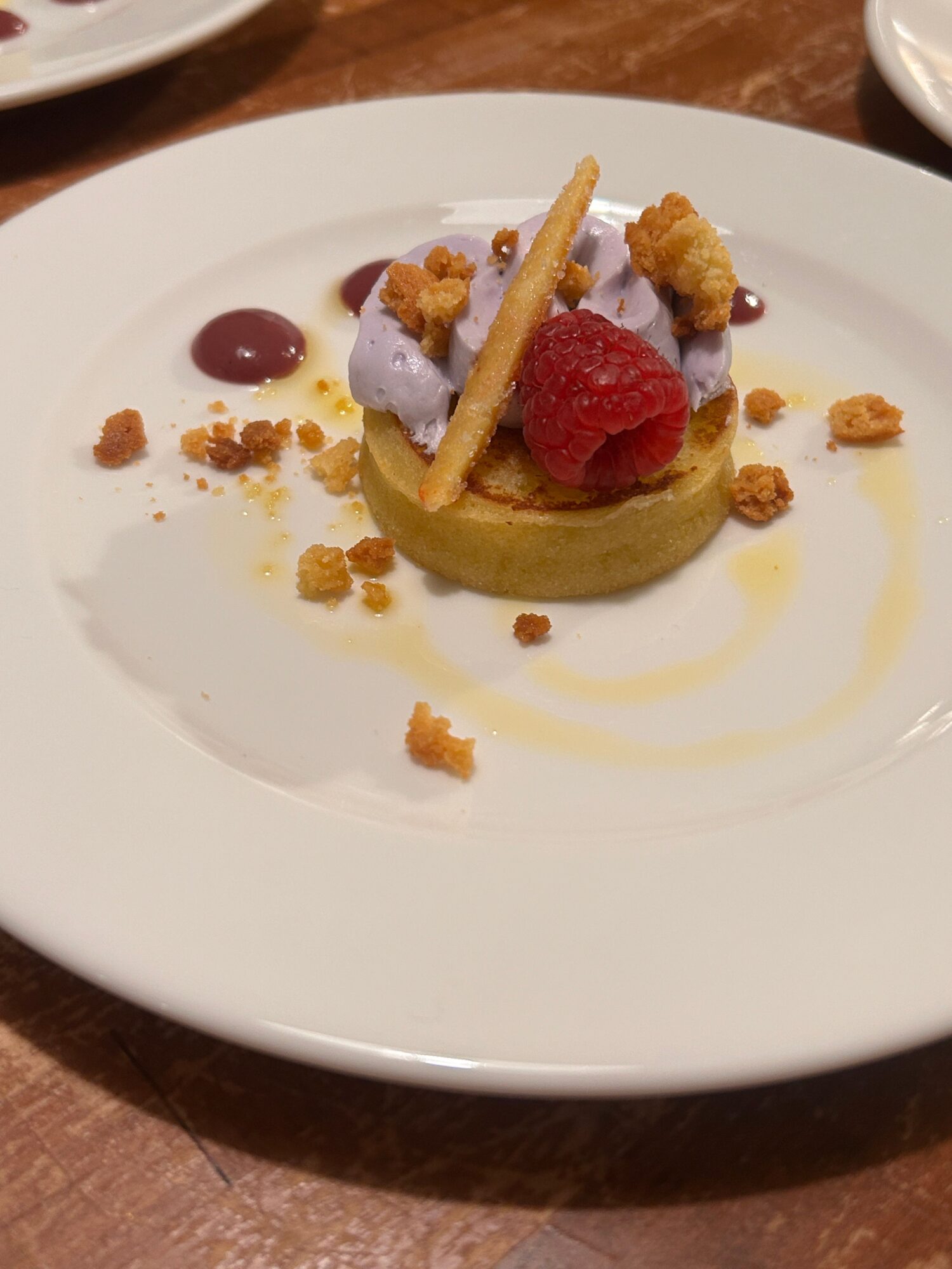 Dessert with raspberry, purple cream, and crunchy topping on a white plate, garnished with sauce and crumbs.