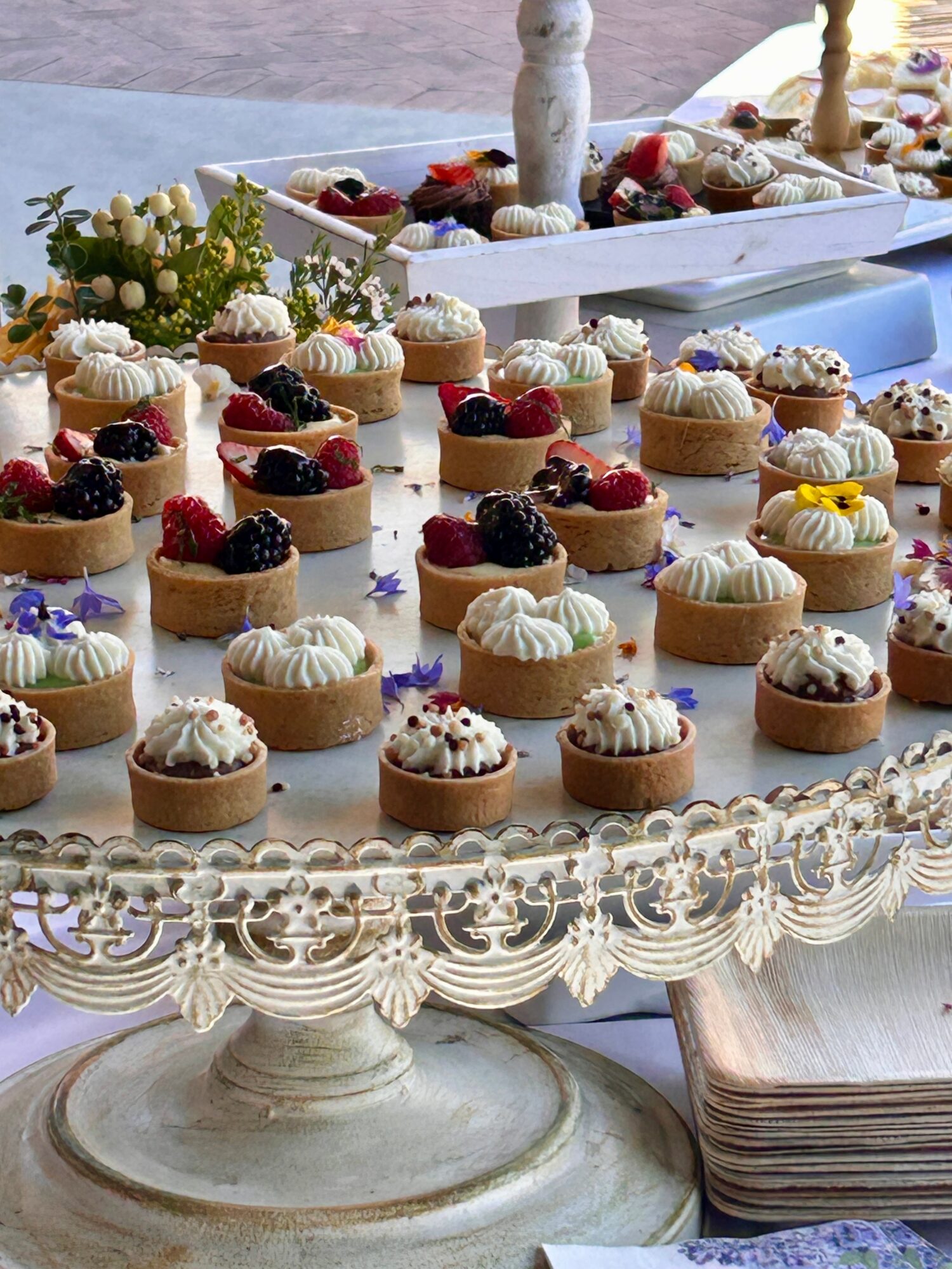Assorted mini desserts with berries and cream on an ornate cake stand, with additional treats in the background.