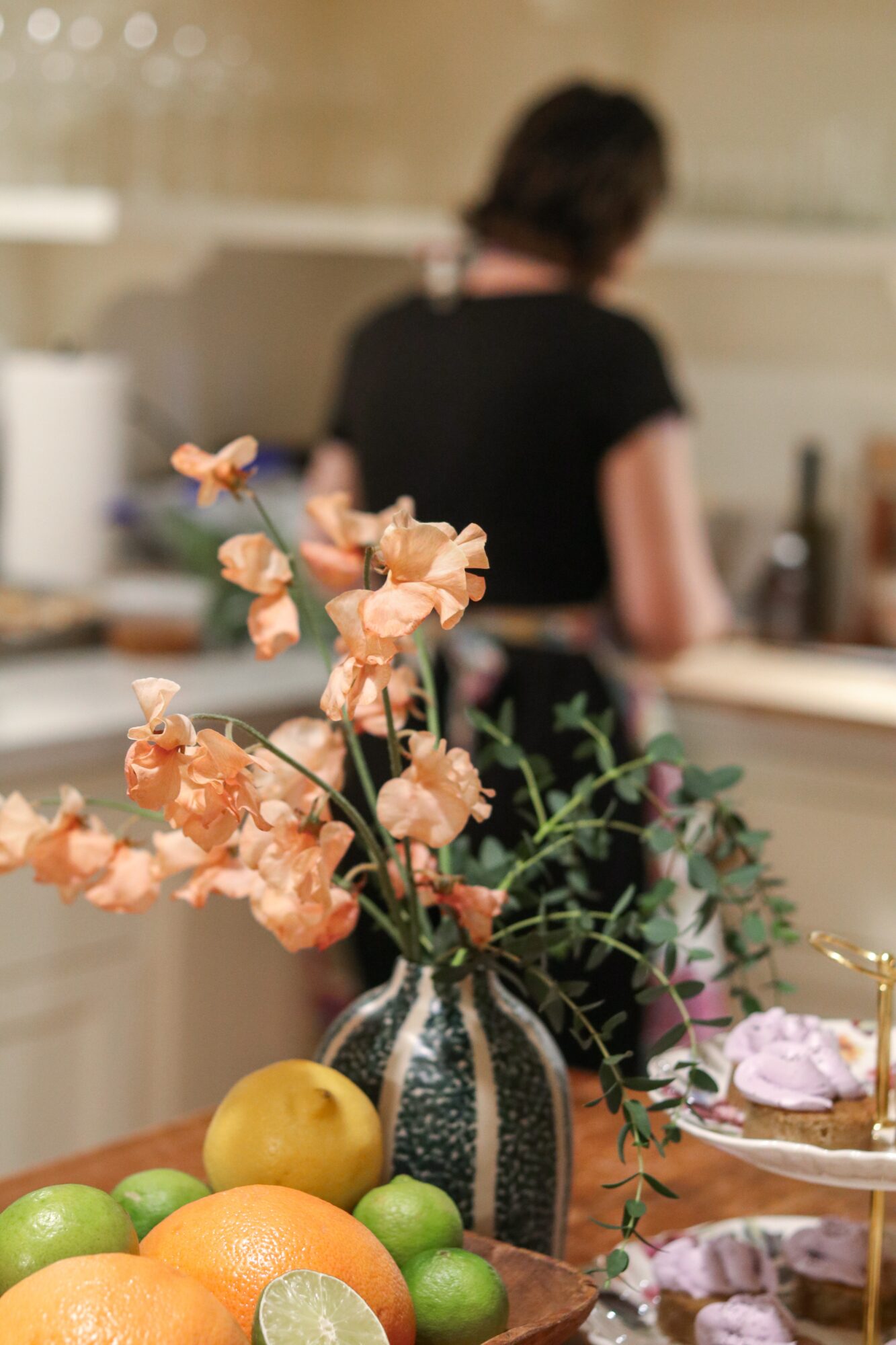 A vase with peach-colored flowers on a table with green and yellow fruits, blurred person in background, kitchen setting.