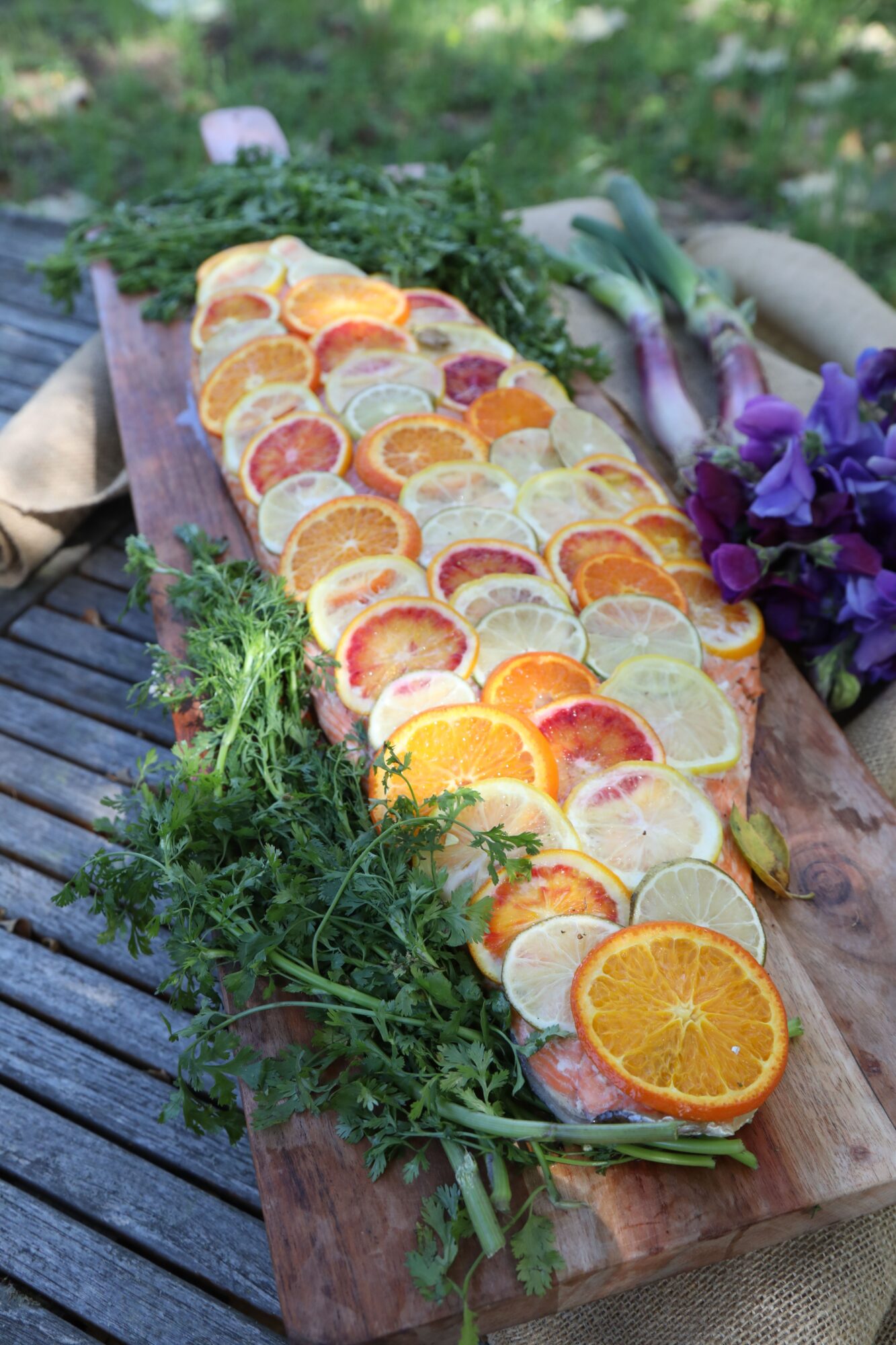 A long wooden board with sliced oranges, lemons, and grapefruits, garnished with green herbs and purple flowers, outdoors.