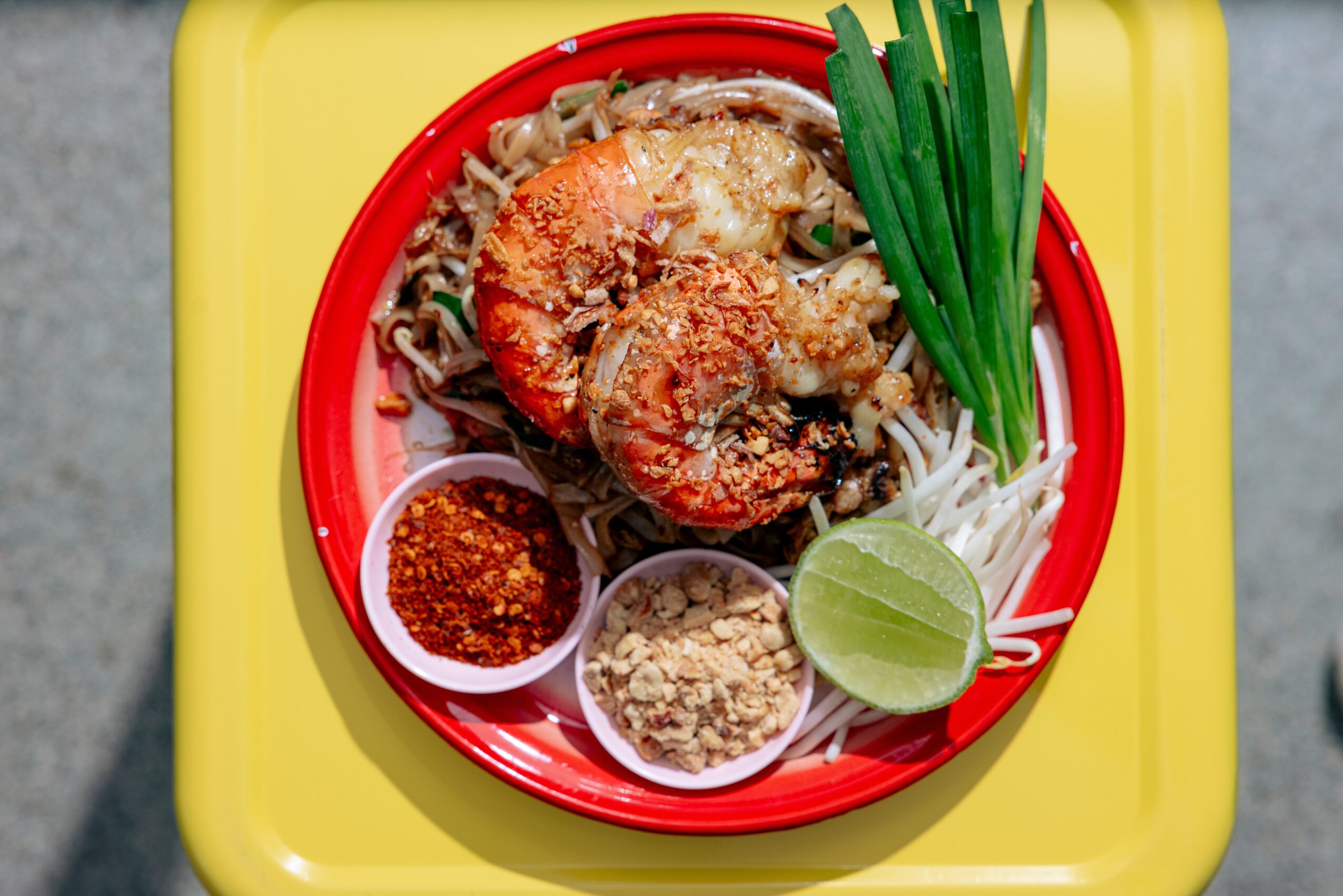 Shrimp on noodles with lime, chili powder, crushed peanuts, and green onions on a yellow tray.