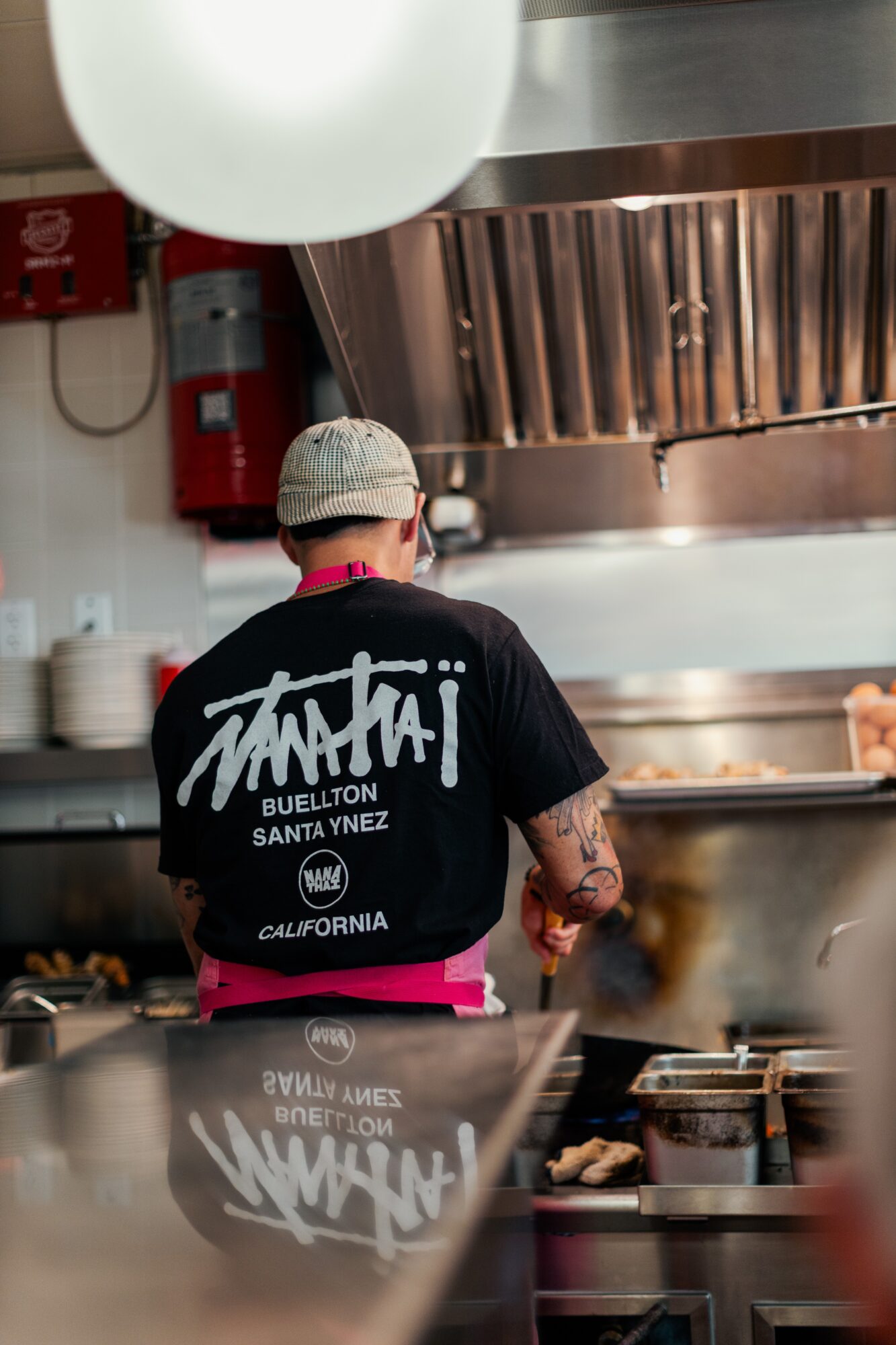 Person wearing a black T-shirt and a cap working in a kitchen with a large metallic vent and fire extinguisher in background.