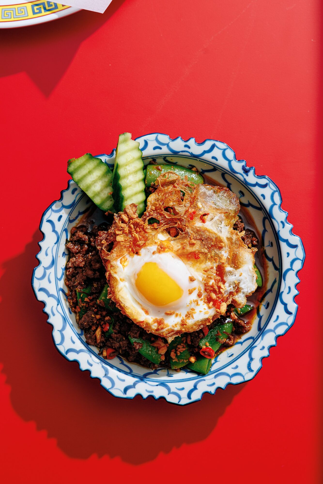 Bowl with fried egg, cucumber slices, and herbs on a red surface.