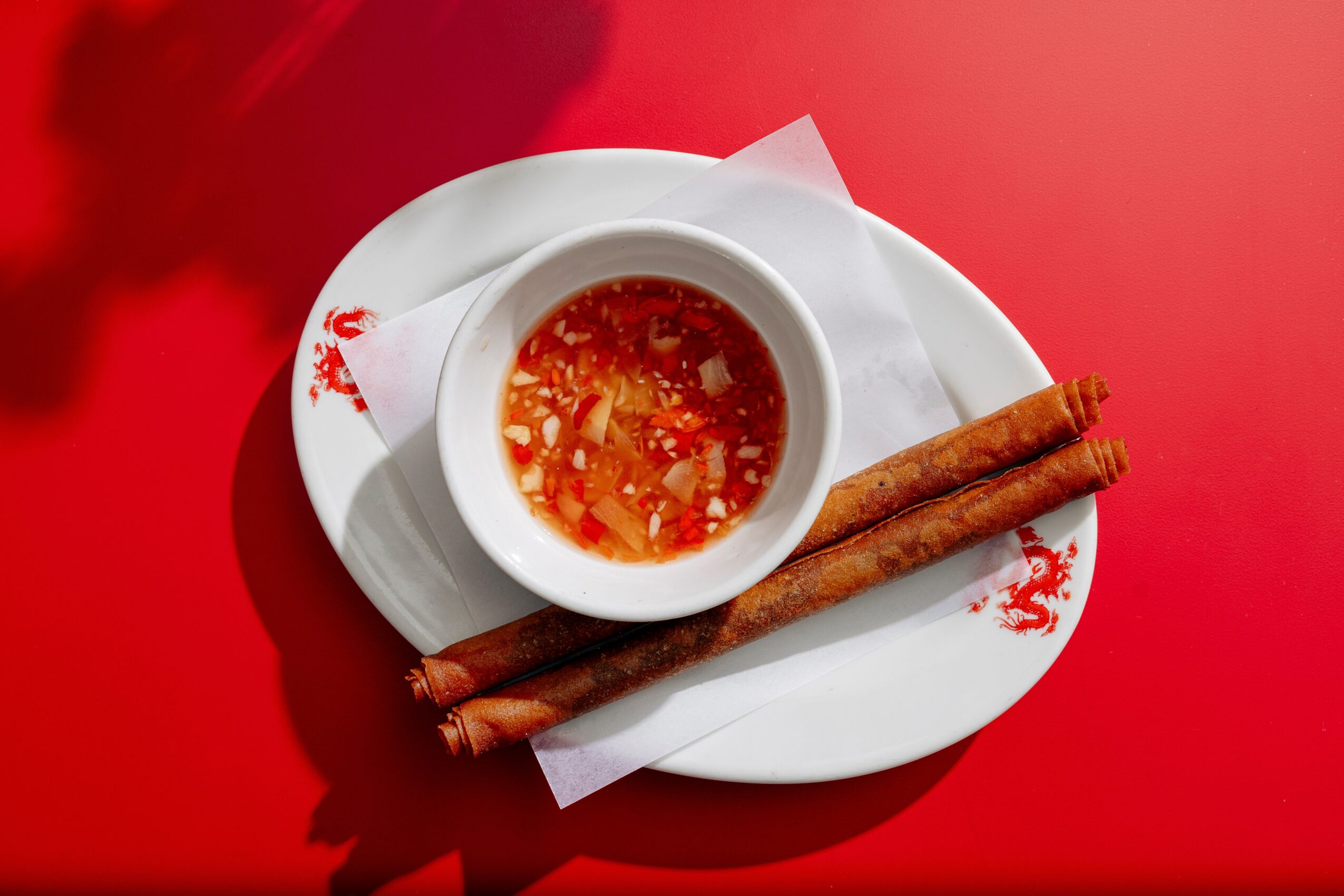 Bowl of dipping sauce with two cinnamon sticks on a white plate, on a red surface.