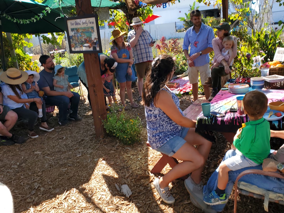 Group of children and adults gathered outdoors around a woman playing a piano, with trees and decorations in the background.