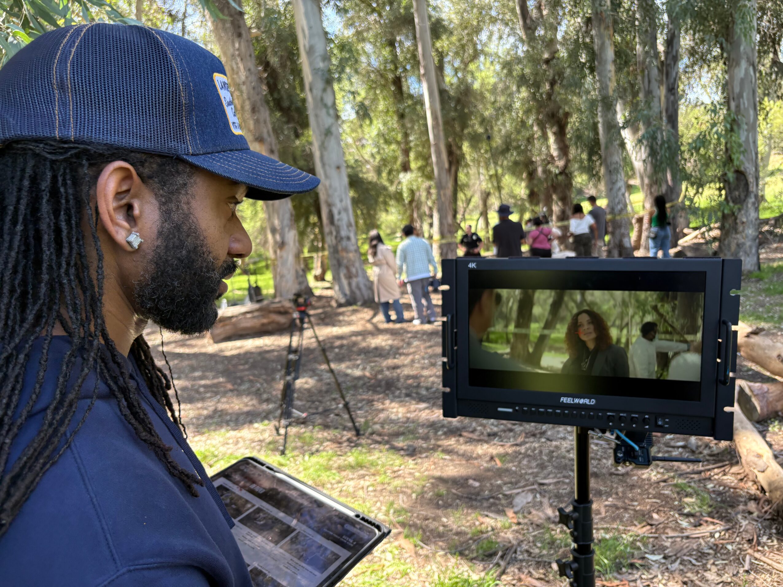 Man with dreadlocks and a cap filming outdoors with a monitor showing a woman, trees in background.