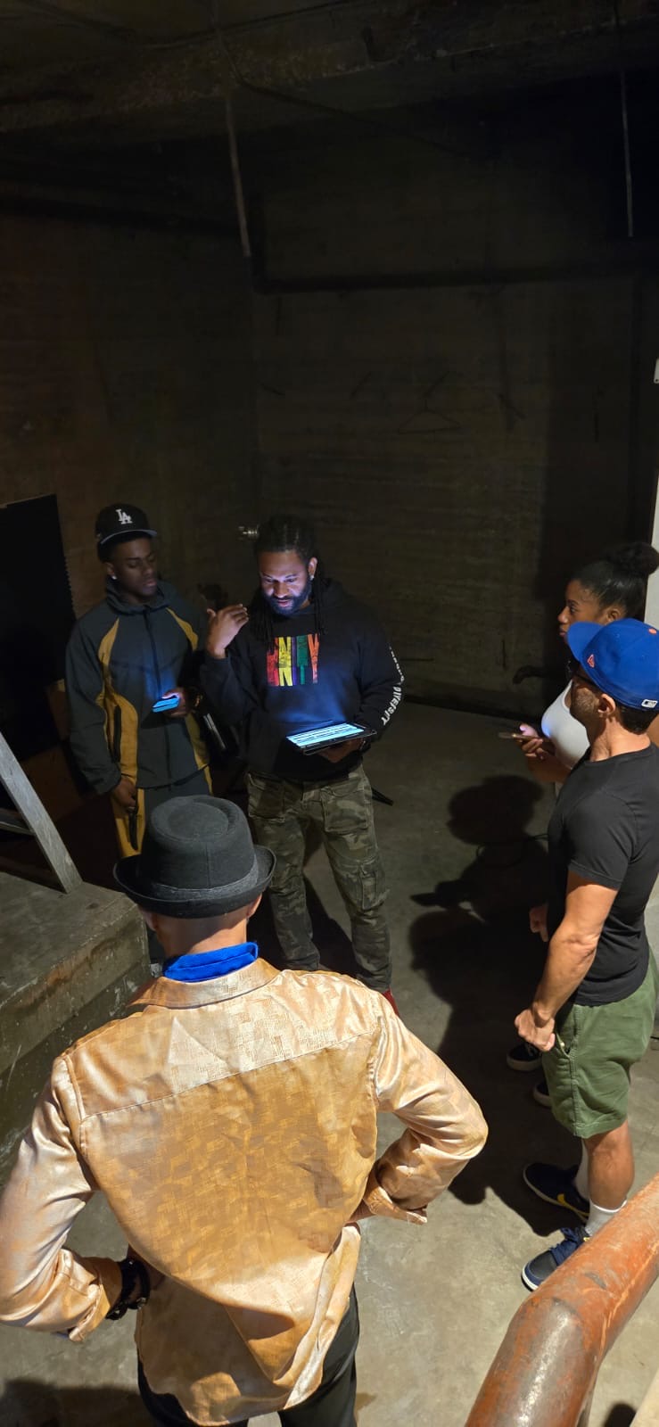 Group of five people standing in a dimly lit indoor space, some wearing hats and masks, engaged in conversation.