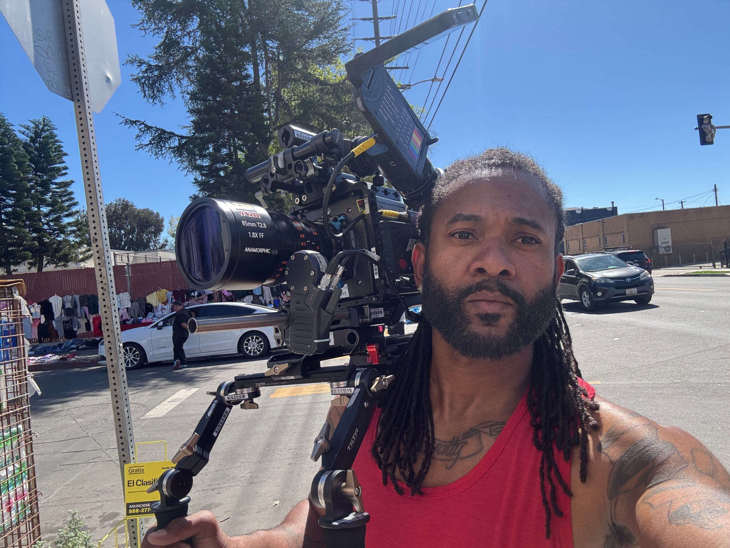 Man with long dreadlocks and a beard wearing a red sleeveless shirt, holding a large professional camera on his shoulder, outdoors on a sunny day.