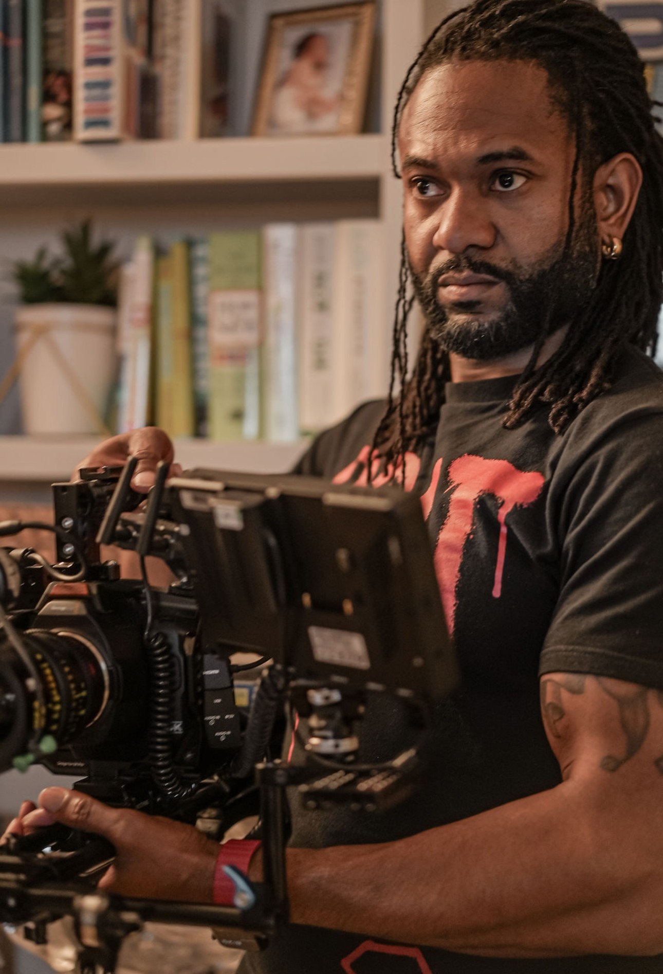 Man with dreadlocks operating professional video camera in indoor setting with books and framed photos in background.