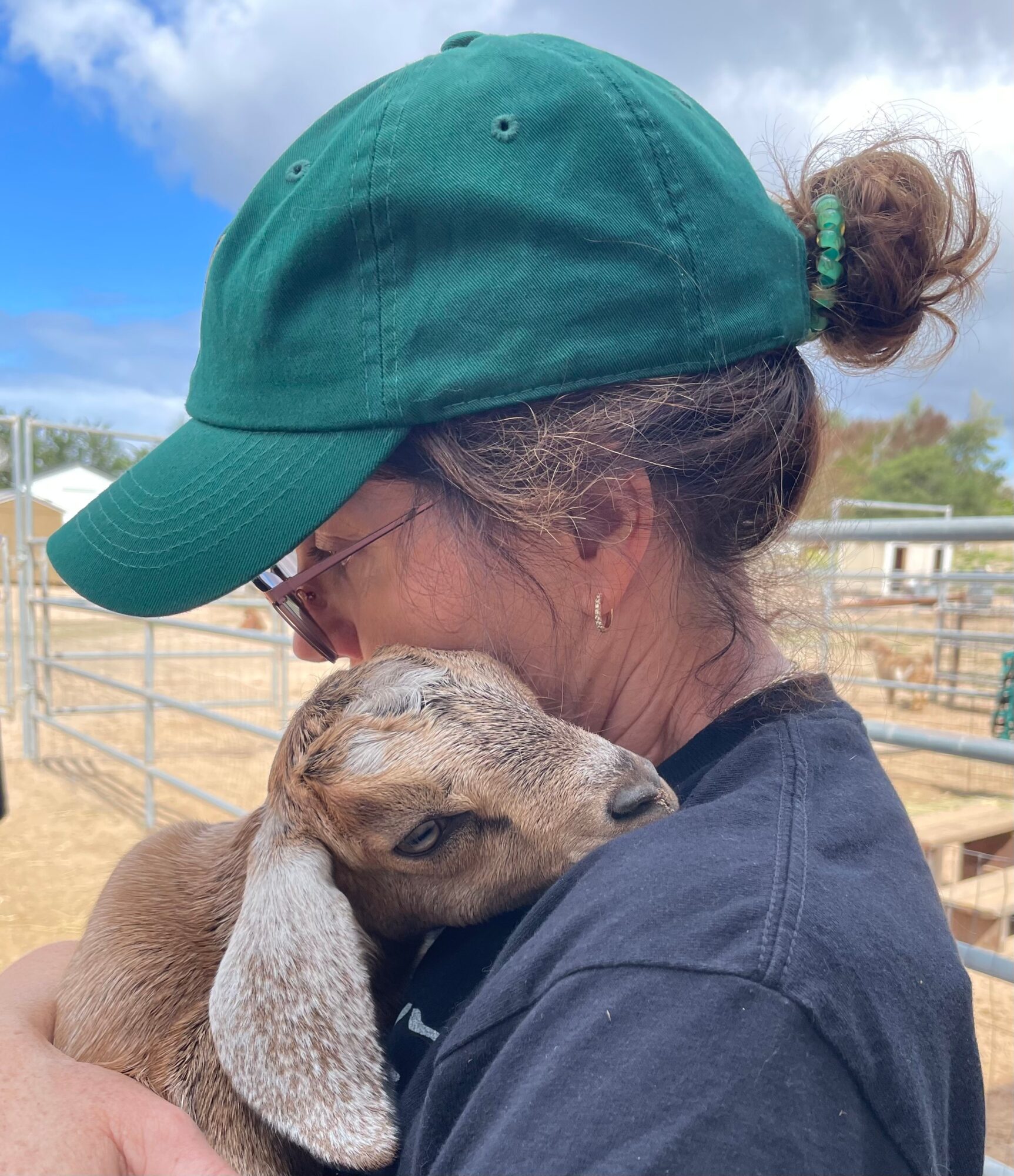 Person wearing a green cap and glasses holding a baby goat outdoors, with a fence and blue sky in background.