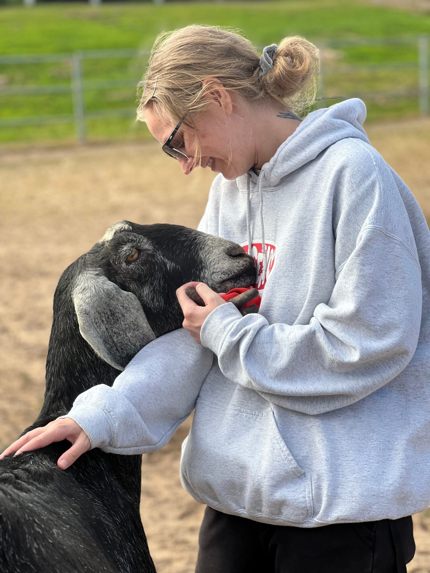 Person with blonde hair in a gray hoodie gently holds a black and white goat's face outdoors.