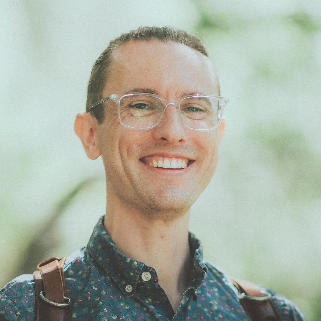 Smiling man with glasses and a patterned shirt outdoors, blurred green background.
