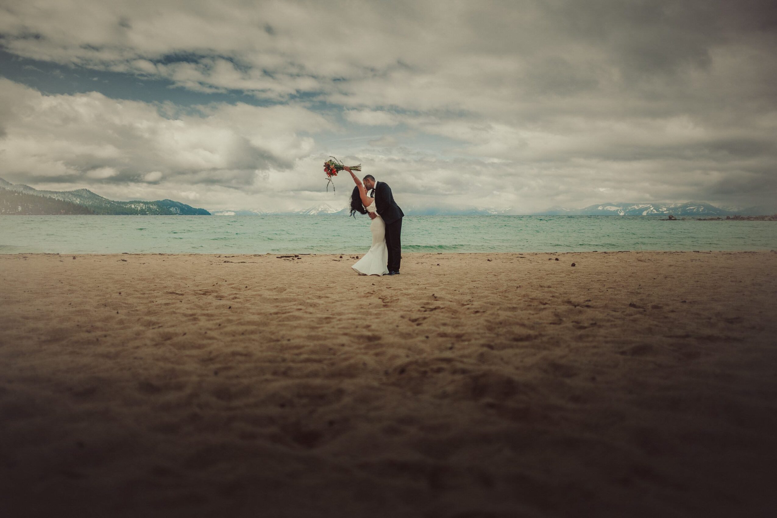 A couple in wedding attire on a beach, with the groom lifting the bride, under cloudy sky and ocean background.