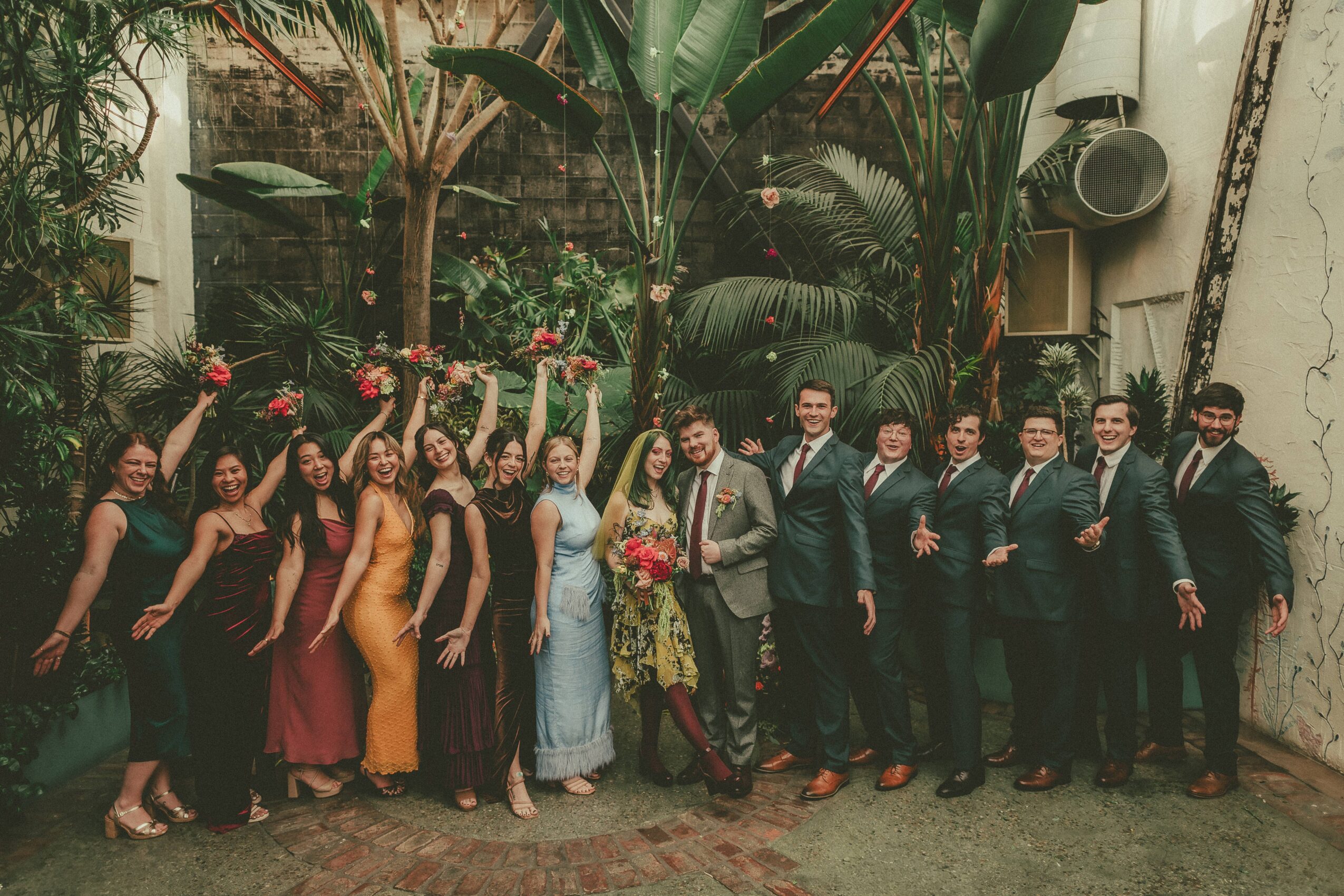 Group of people dressed in formal attire, standing outdoors with plants and trees in the background.