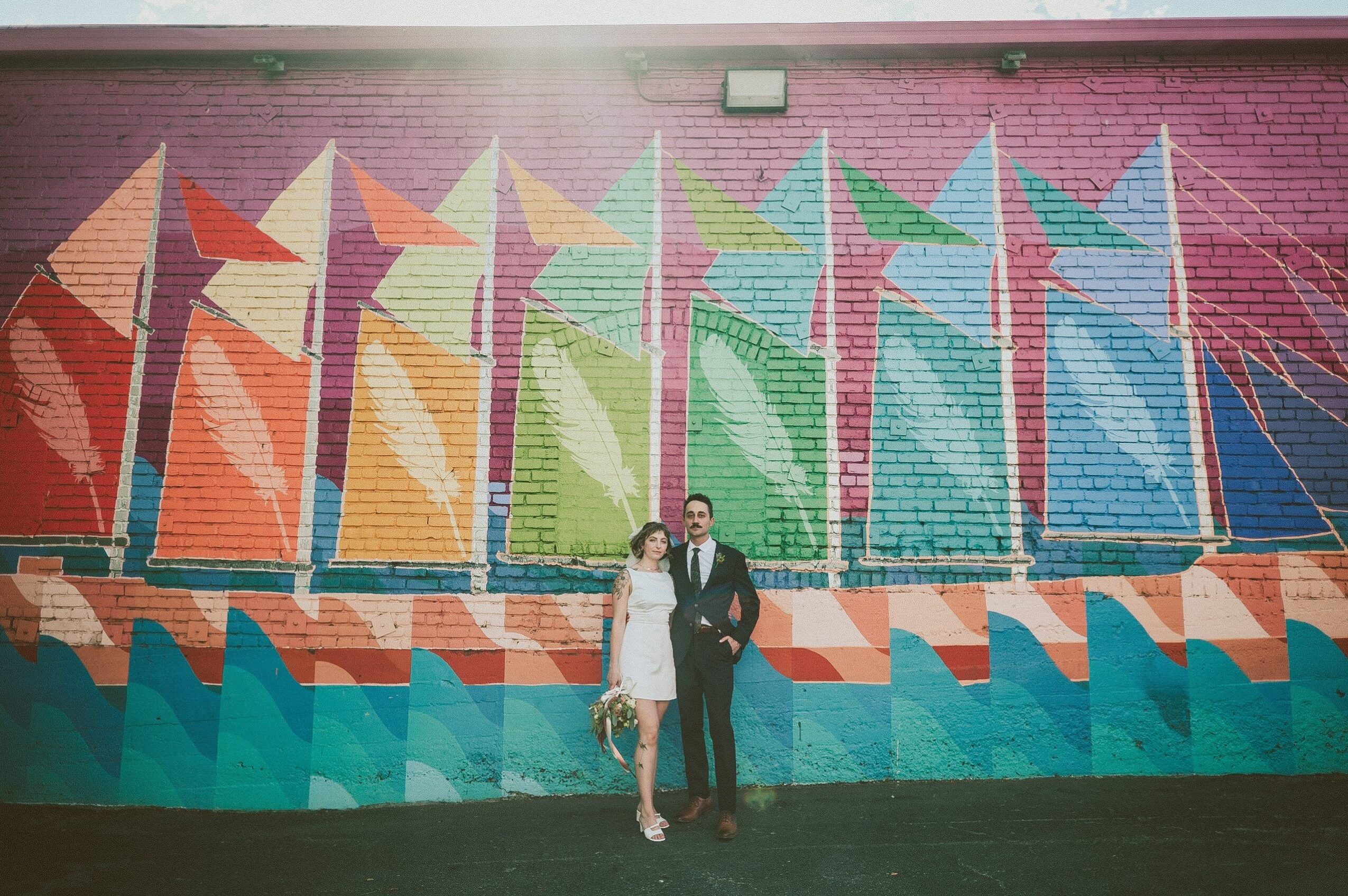 Two people stand in front of a colorful mural with geometric shapes and patterns on a brick wall.