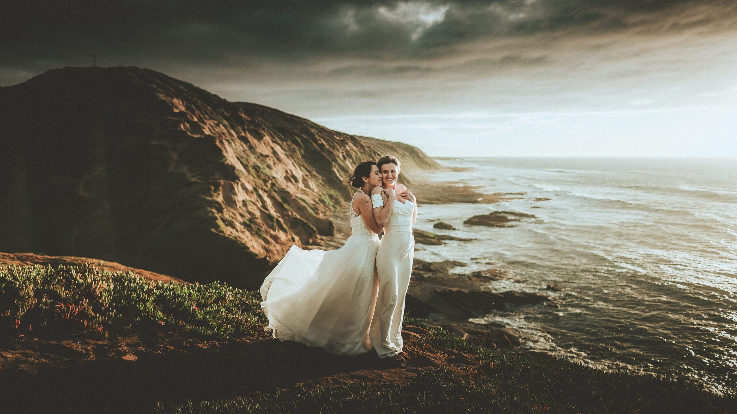 A couple in wedding attire stands on a cliff overlooking the ocean at sunset, with a dramatic sky and rugged coastline.