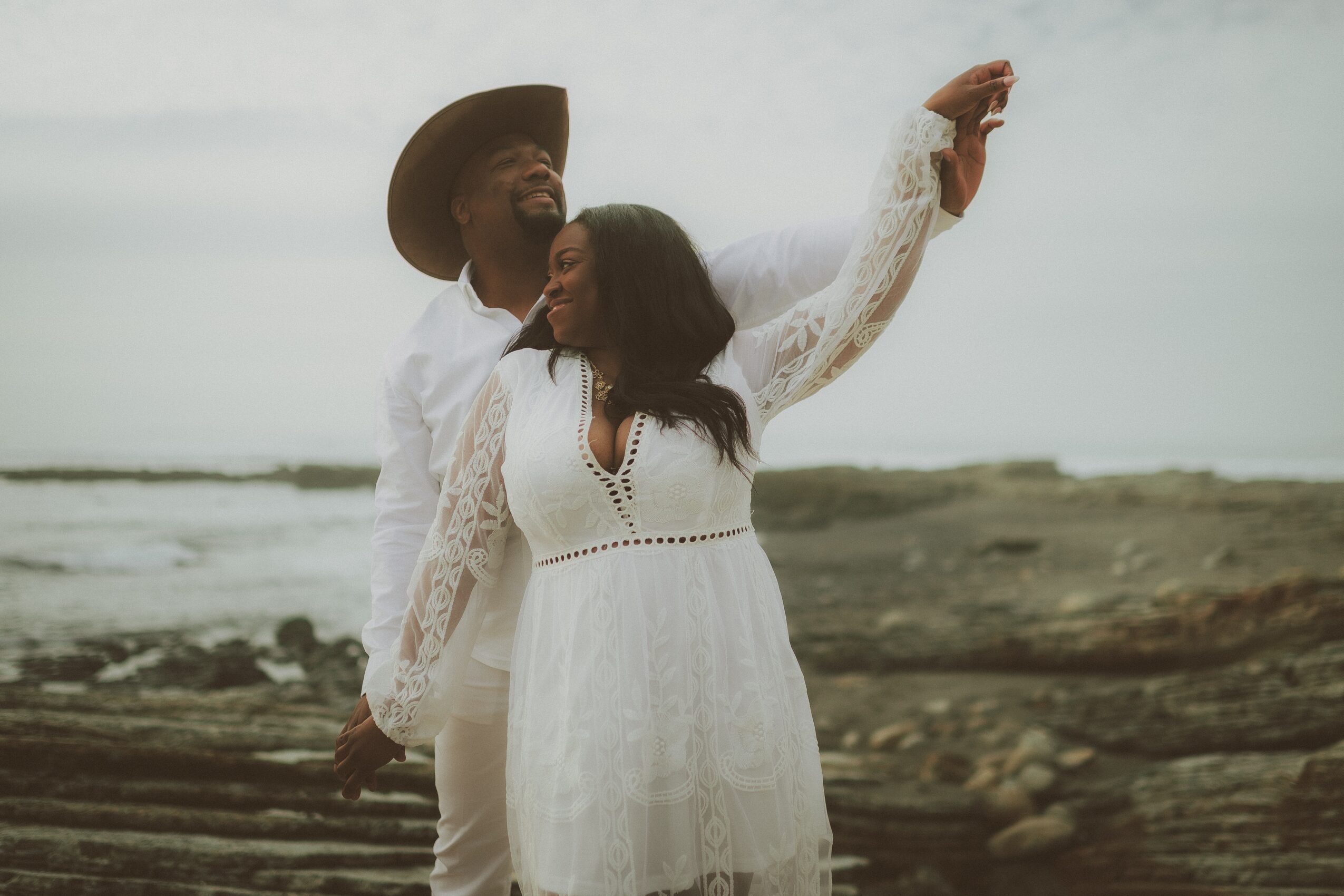 A couple stands on a rocky shoreline, with the woman raising her arm and the man wearing a hat, both smiling.