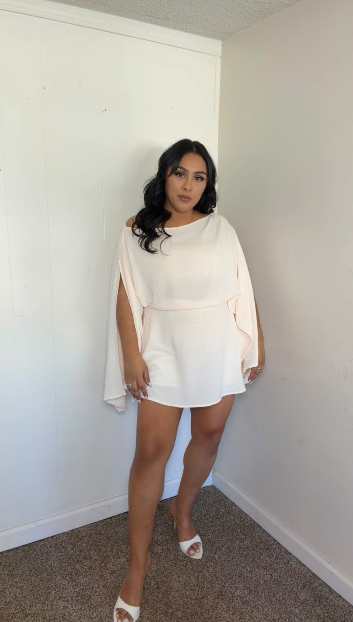 Woman with dark hair in a white dress standing in a room with white walls and carpeted floor.