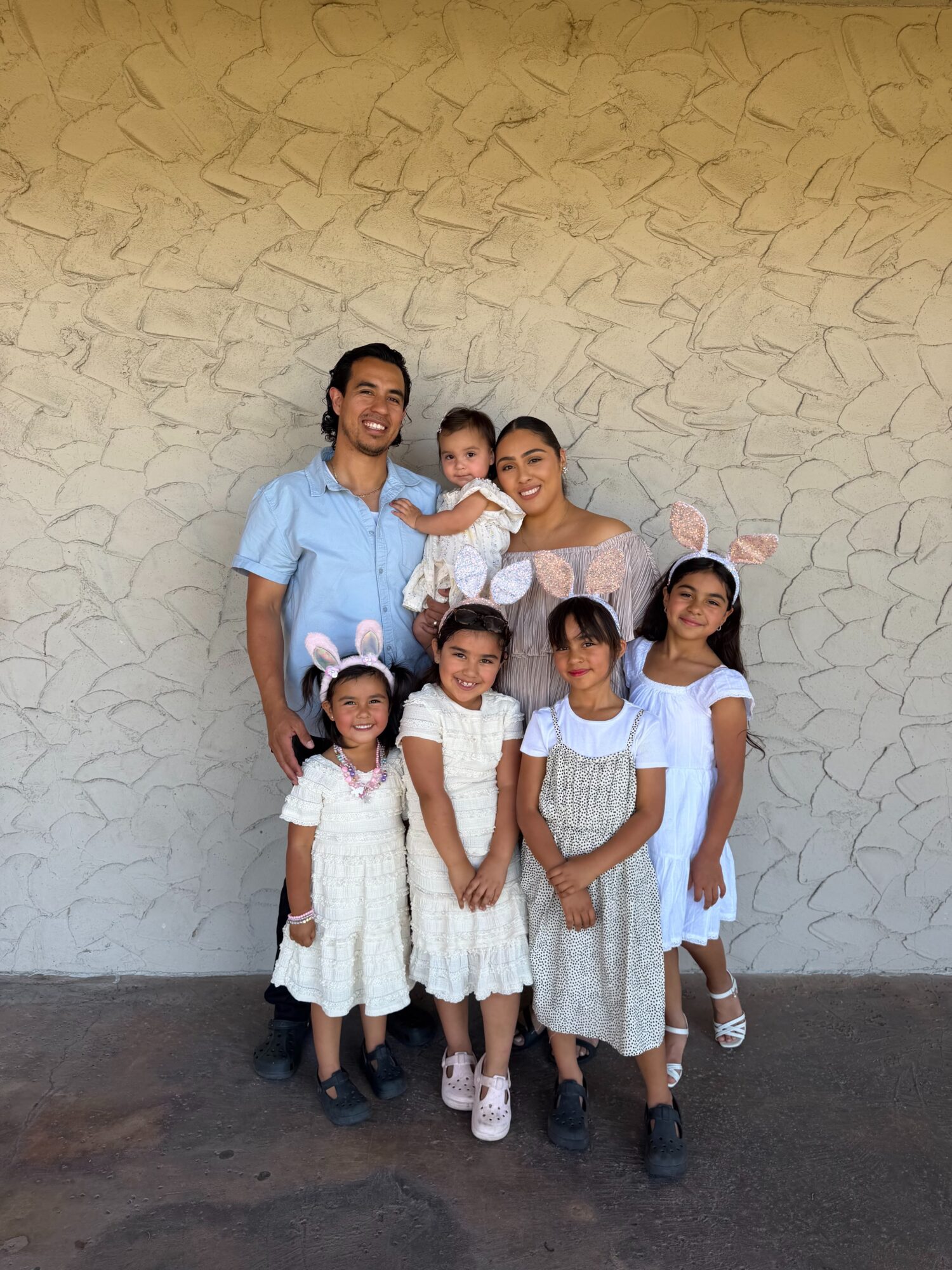 Group of six people, including children and adults, standing against a textured wall, smiling, with one child being held.